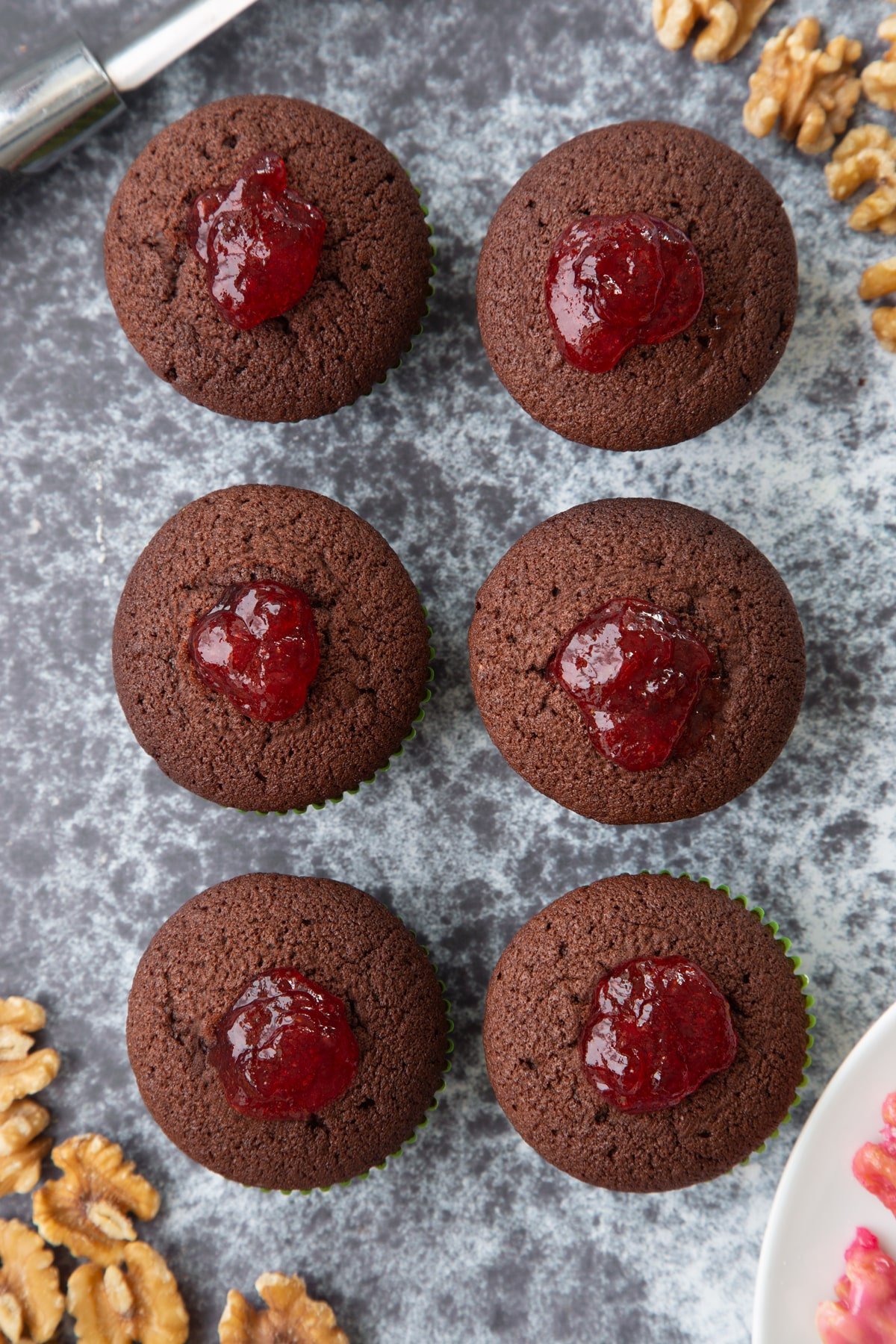 Six chocolate cupcakes filled with strawberry jam. Ingredients to make gory Halloween cupcakes surround them.