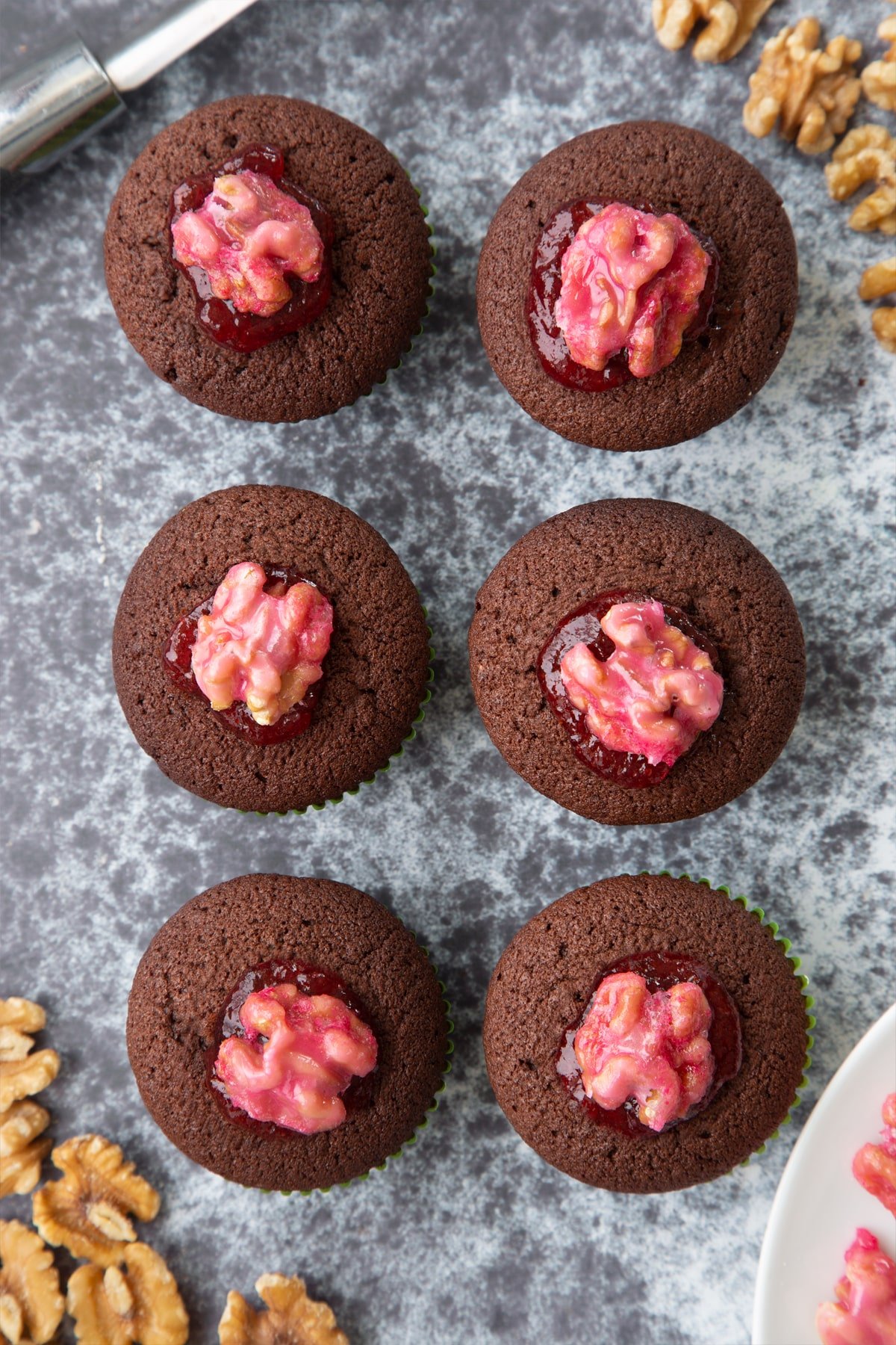 Gory Halloween cupcakes on a black background from above. The cupcakes are filled with jam and topped with pink candied walnuts that look like brains.