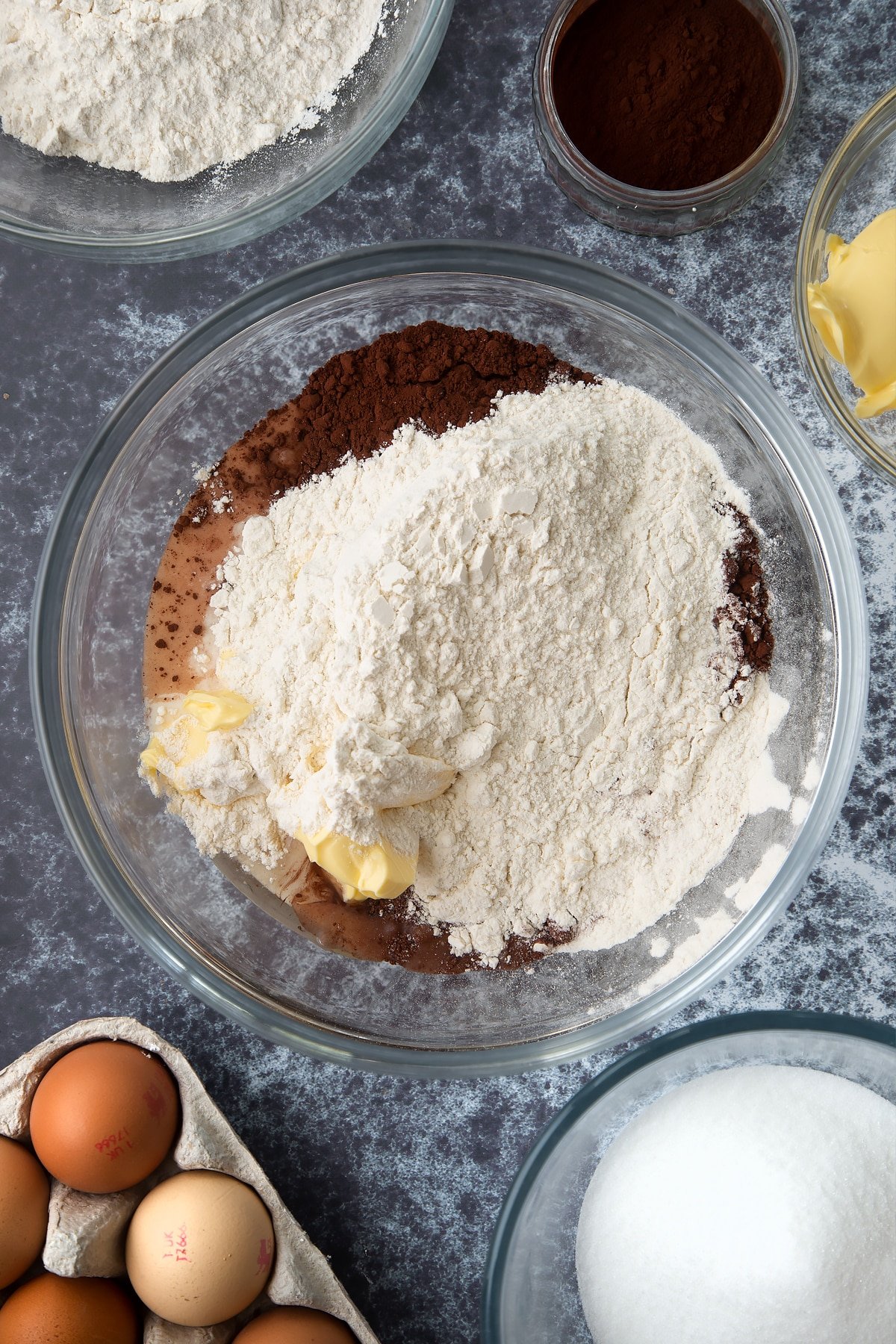 Margarine, oil, caster sugar, milk, cocoa, eggs and self-raising flour in a large mixing bowl. Ingredients to make gory Halloween cupcakes surround the bowl.