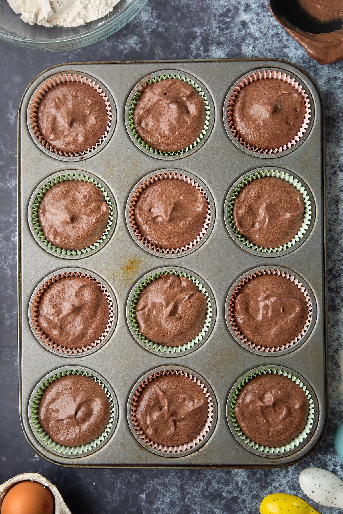 Chocolate cupcake batter in a lined 12-hole muffin tray. Ingredients to make gory Halloween cupcakes surround the bowl.