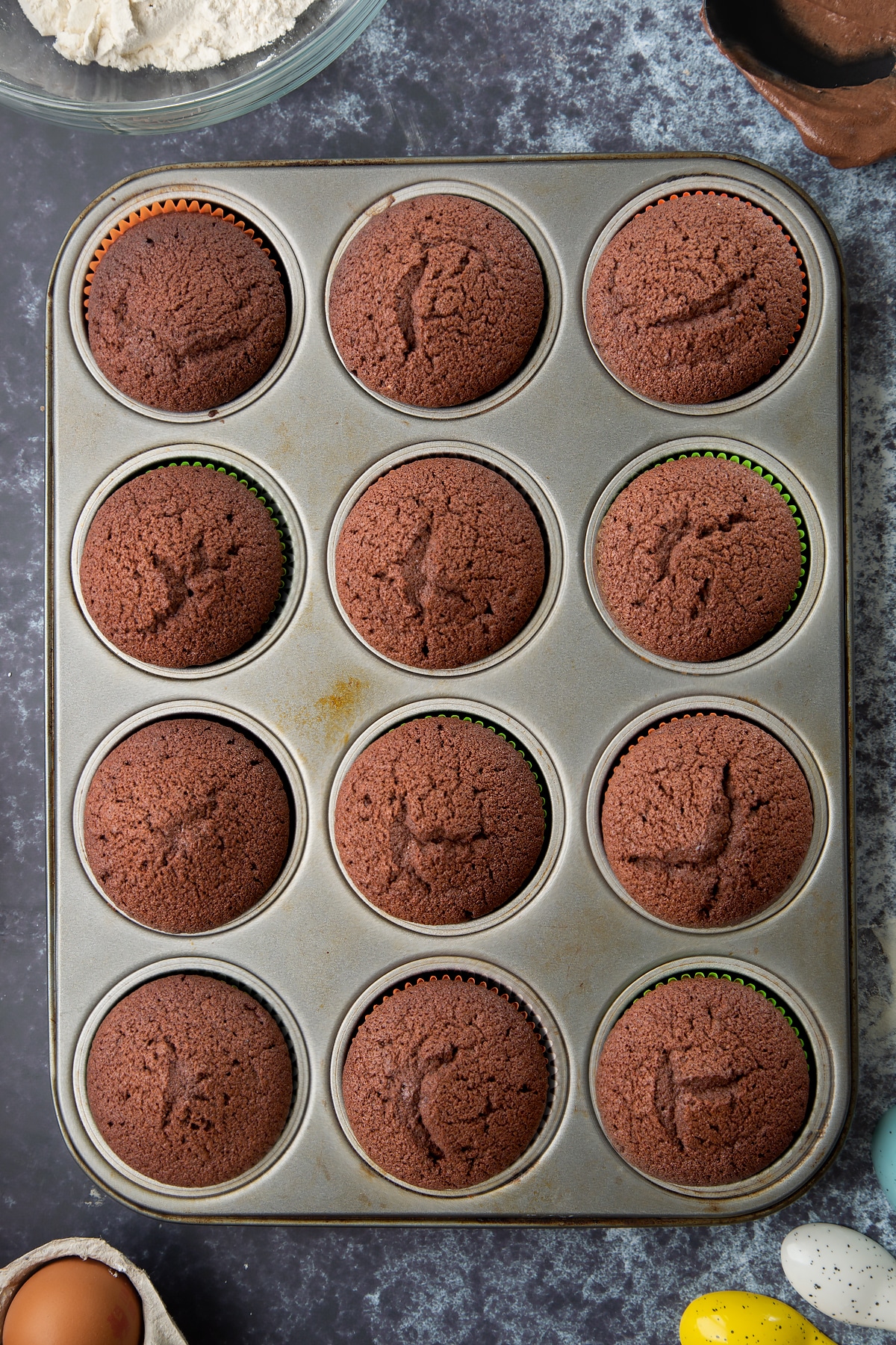 Chocolate cupcakes in a lined 12-hole muffin tray. Ingredients to make gory Halloween cupcakes surround the bowl.