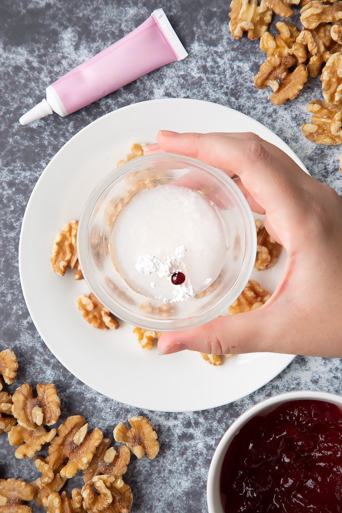 Walnuts on a white plate. Above, a hand holds a bowl containing icing sugar, water and pink food colouring. Ingredients to make gory Halloween cupcakes surround them.