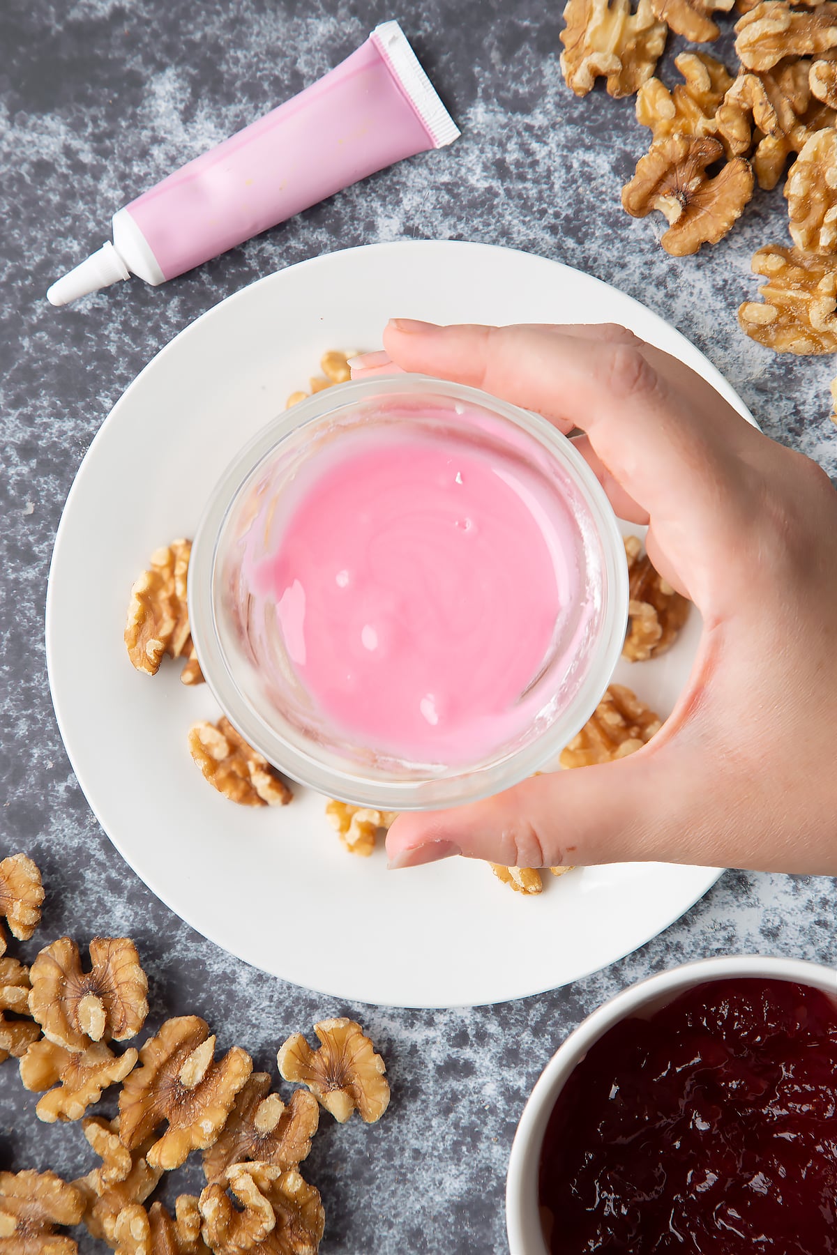 Walnuts on a white plate. Above, a hand holds a bowl containing pink icing. Ingredients to make gory Halloween cupcakes surround them.