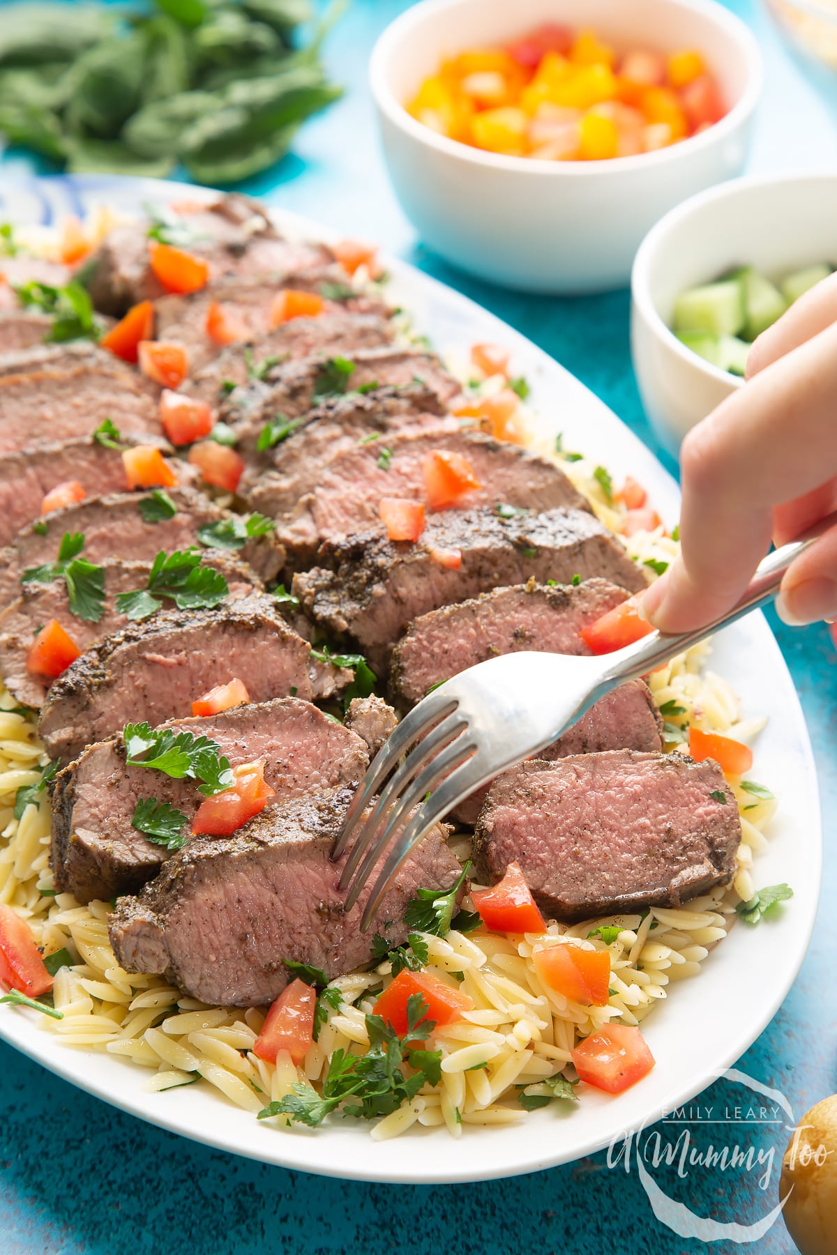 Slices of herby lamb arranged on a bed of orzo on an oval platter. A hand with a fork delves in.