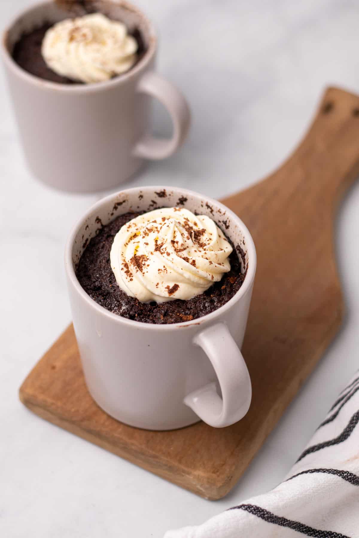 Chocolate mug cakes. One mug sits on a wooden board. The mug cakes have been topped with s swirl of cream.