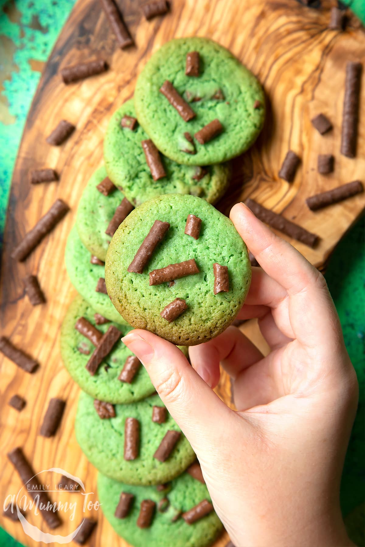 Hand holding a green cookie topped with Matchmaker pieces. More Matchmaker cookies are arranged on a wooden board below.