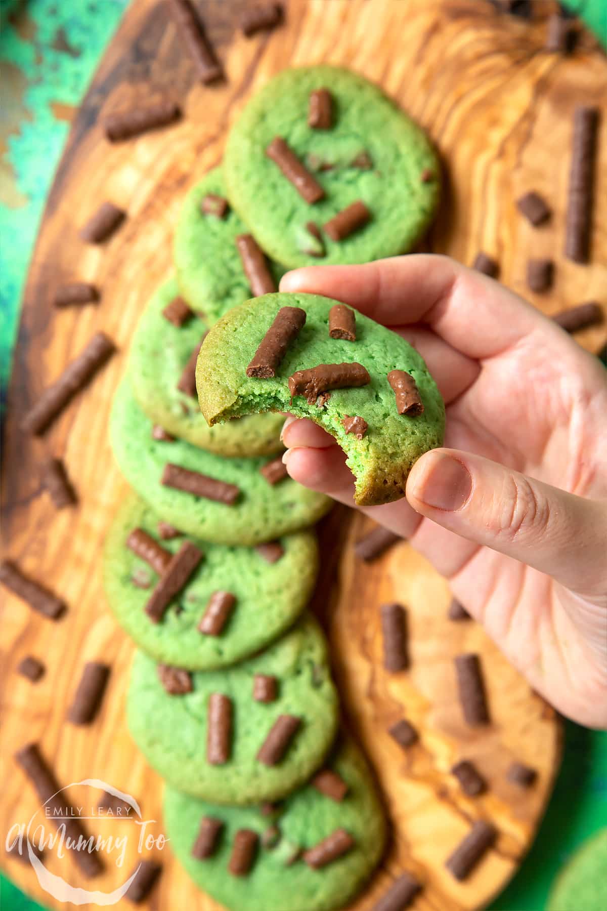 Hand holding a green cookie with a bite out of it. The cookie is topped with Matchmaker pieces. More Matchmaker cookies are arranged on a wooden board below.