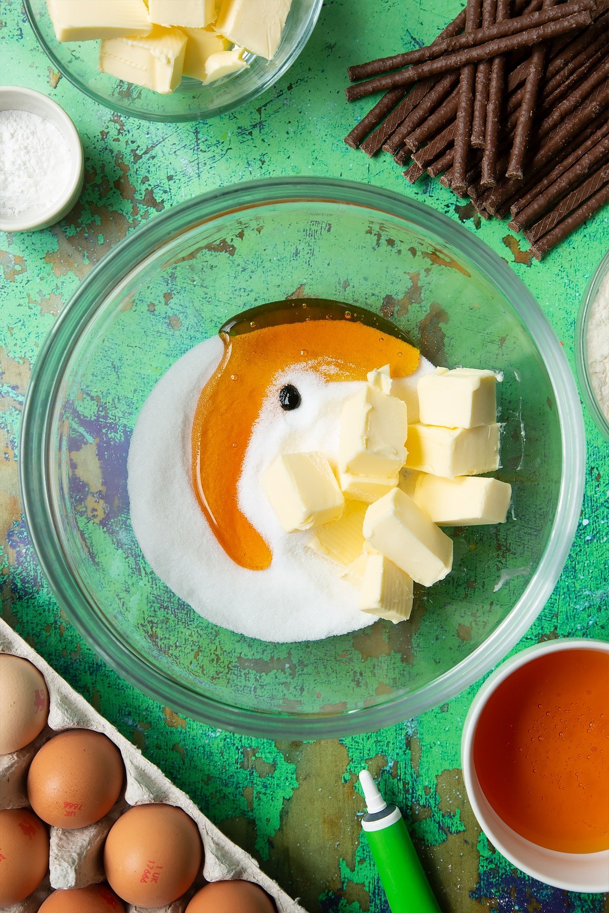 Butter, sugar, golden syrup and green food colouring in a mixing bowl. Ingredients to make Matchmaker cookies surround the bowl.