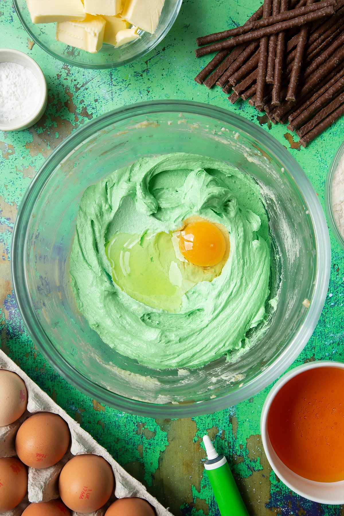 Creamed butter, sugar and green food colouring topped with an egg in a mixing bowl. Ingredients to make Matchmaker cookies surround the bowl.