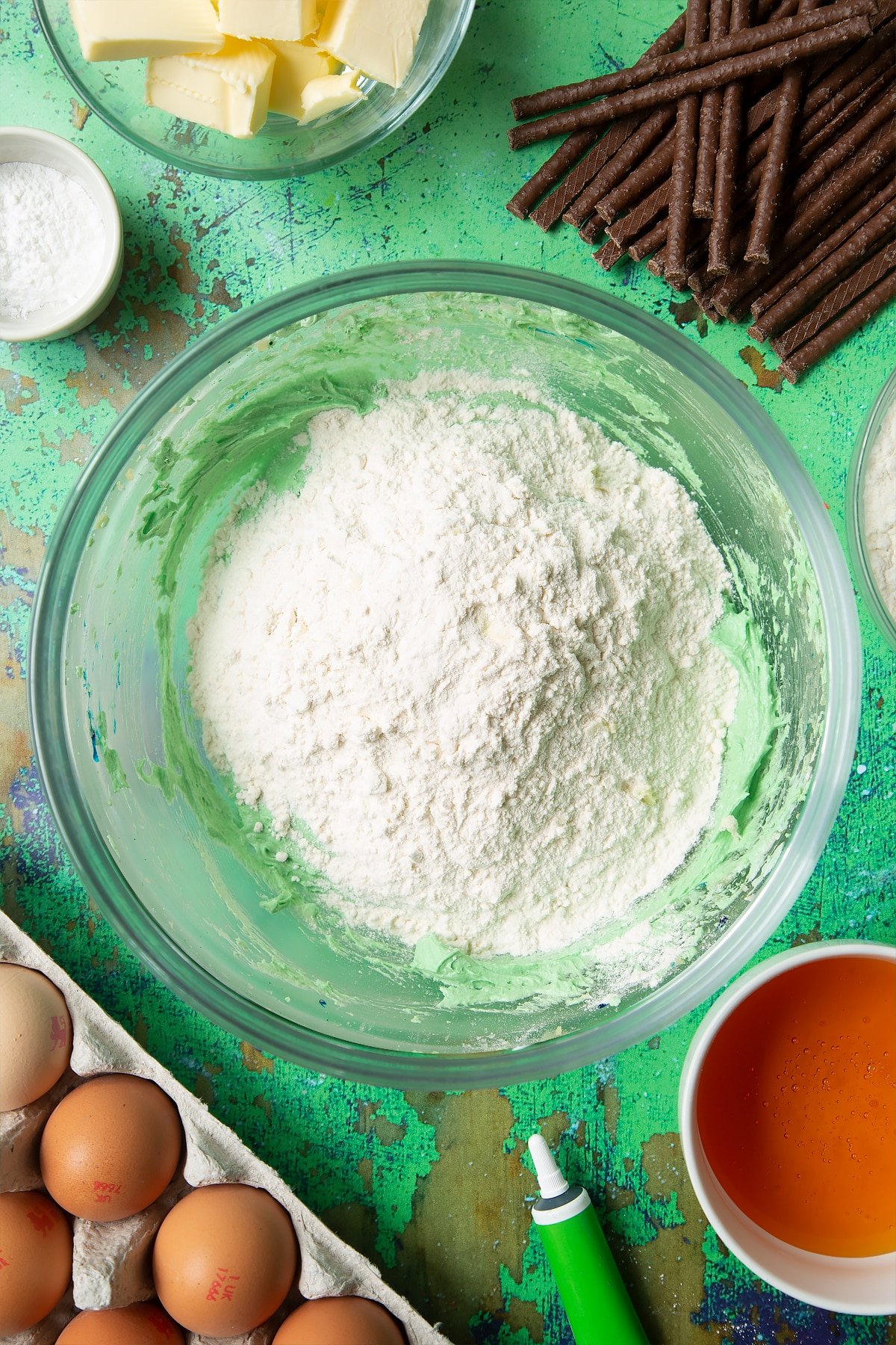 Creamed butter, sugar, eggs and green food colouring topped with flour in a mixing bowl. Ingredients to make Matchmaker cookies surround the bowl.