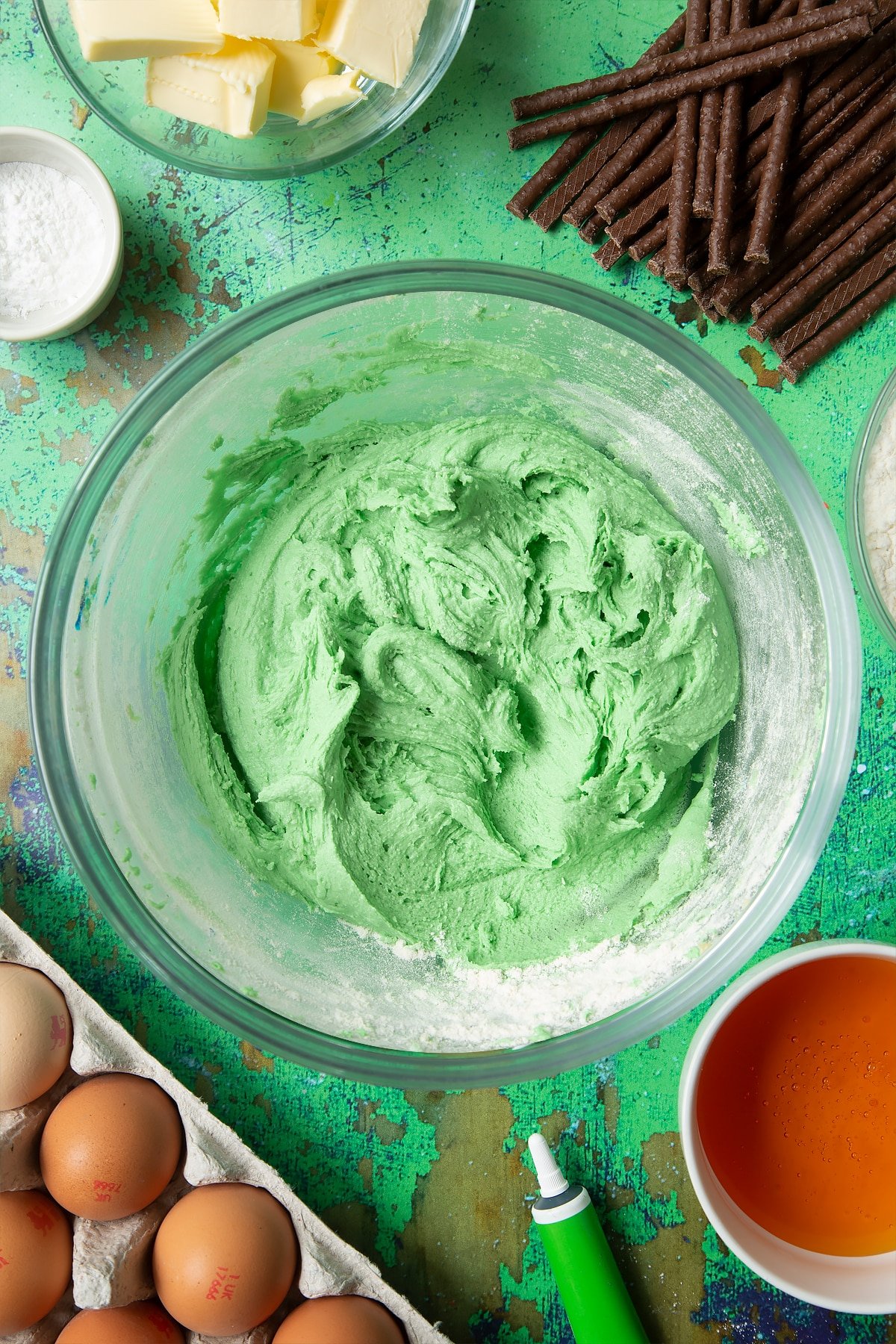 Green cookie dough in a mixing bowl. Ingredients to make Matchmaker cookies surround the bowl.