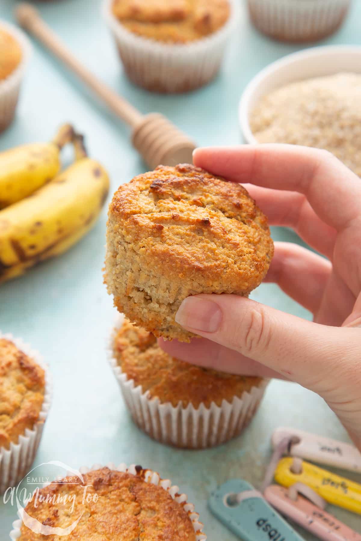 Front angle shot of a hand holding an Oat bran muffin with a mummy too logo in the lower-left corner