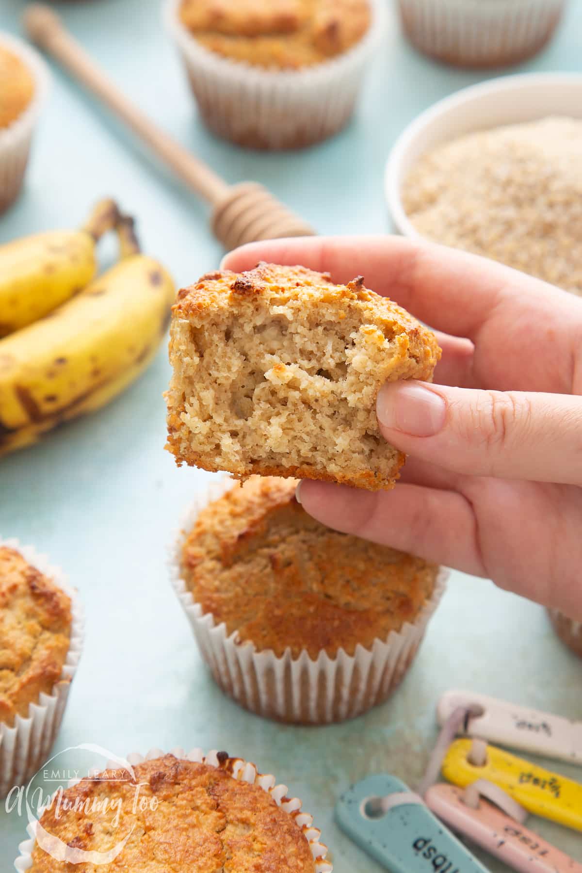 front angle shot of a hand holding a half eaten breakfast oat granola muffin with a mummy too logo in the lower-left corner 