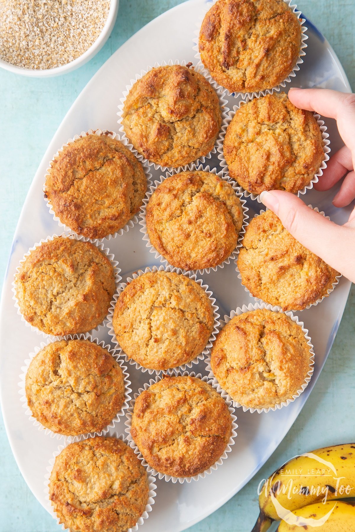 Overhead shot of a hand touching an oat bran granola muffin on a white plate with a mummy too logo in the lower-left corner