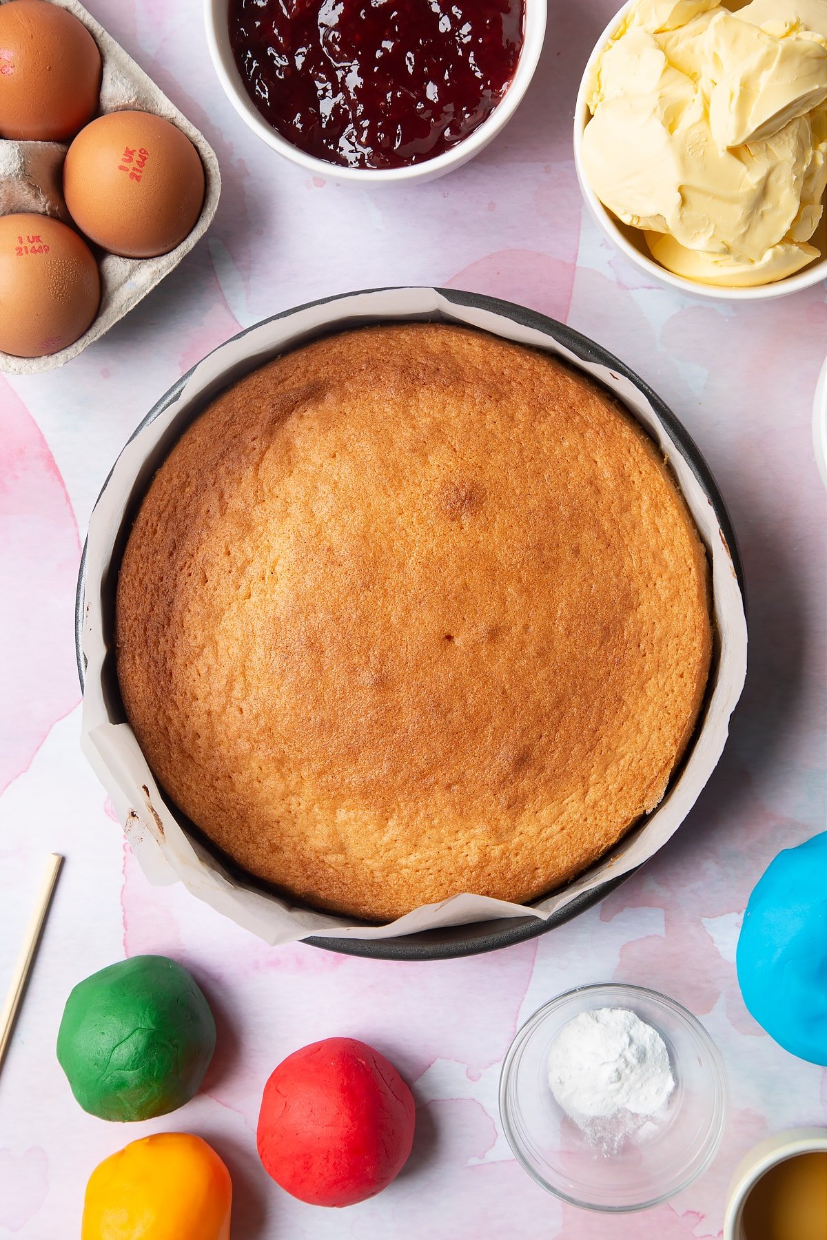 Overhead shot of the baked cake inside a cake tin surrounded by some of the ingredients required to make the sail boat cake. 