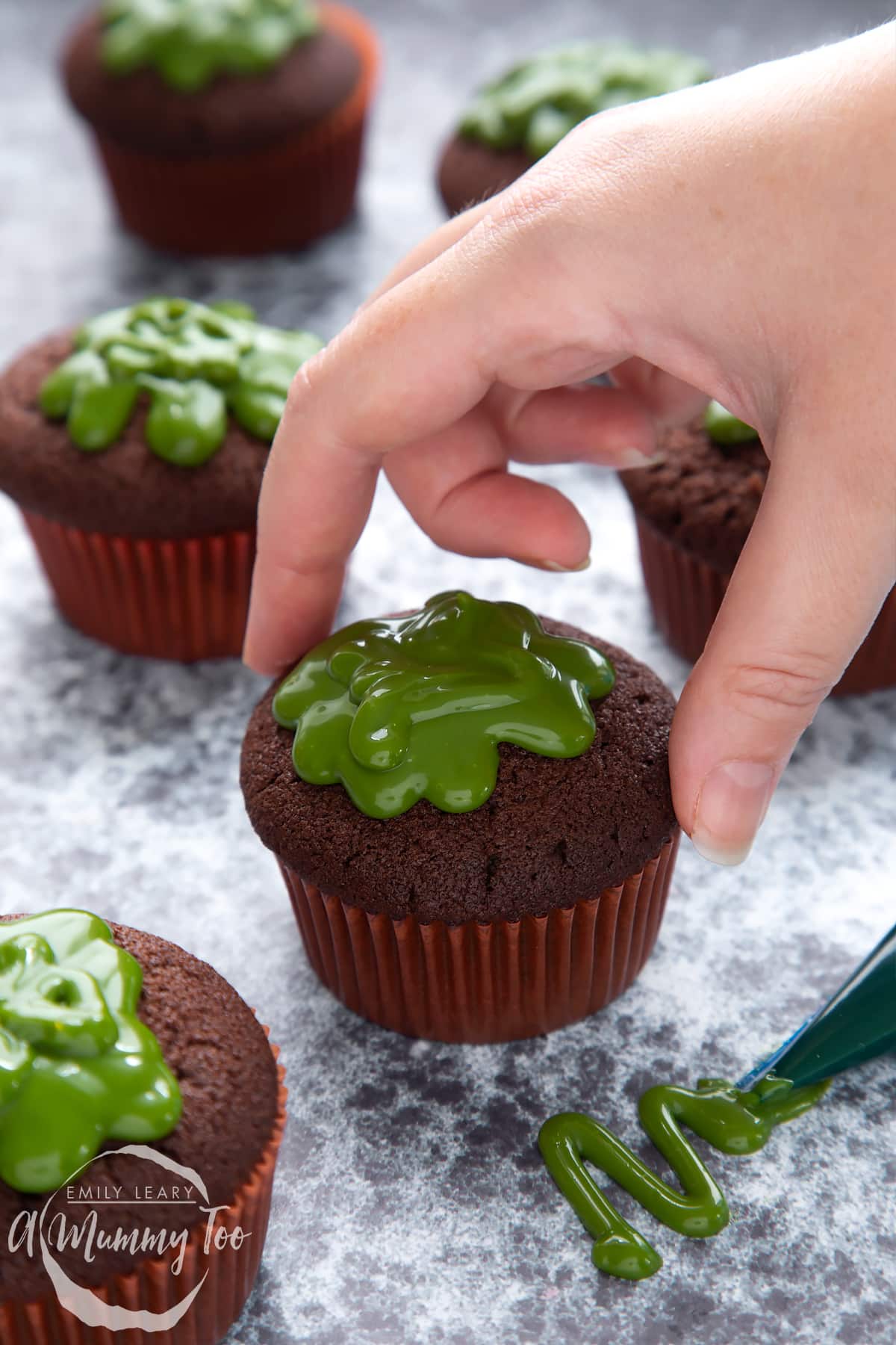 Slime cupcakes on a black backdrop. The cakes have a chocolate sponge topped with dyed-green caramel. A hand reaches to take one.