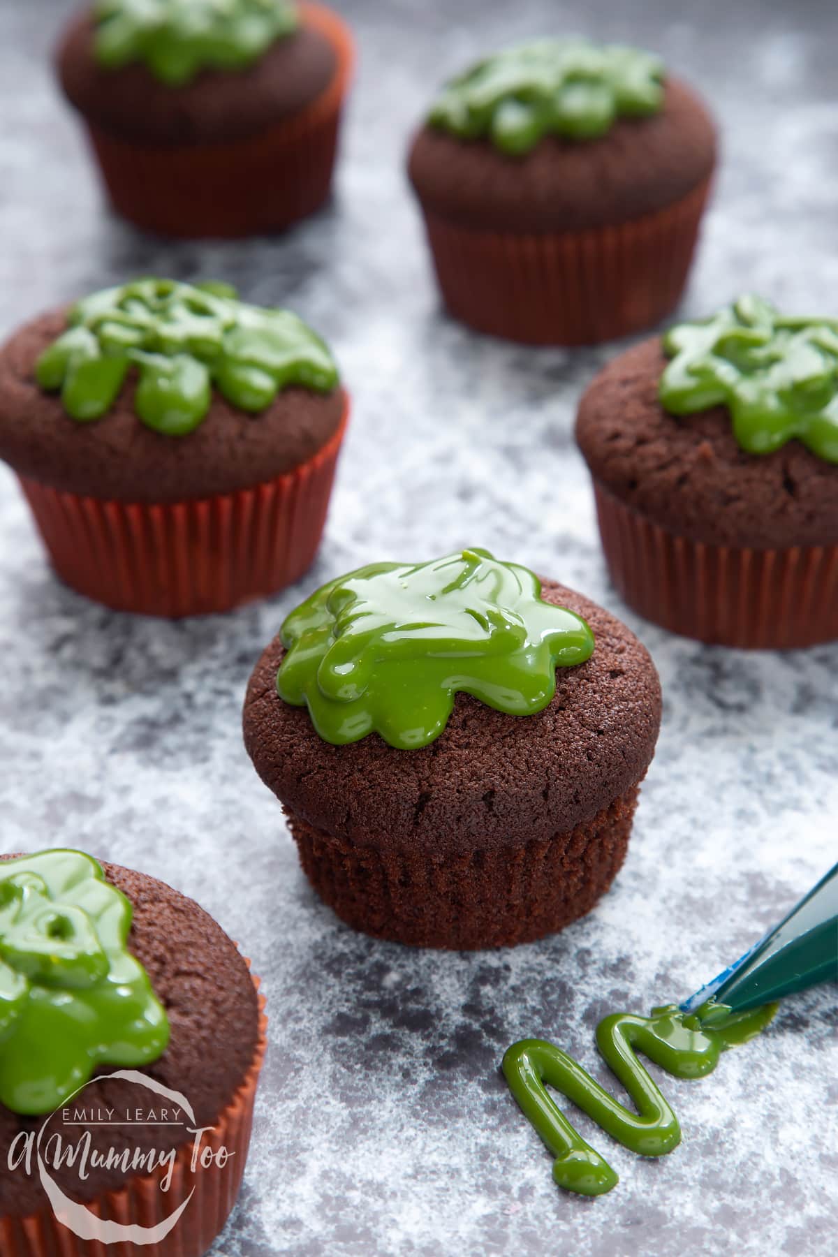 Slime cupcakes on a black backdrop. The cakes have a chocolate sponge topped with dyed-green caramel. The case has been removed from the cupcake in the foreground.