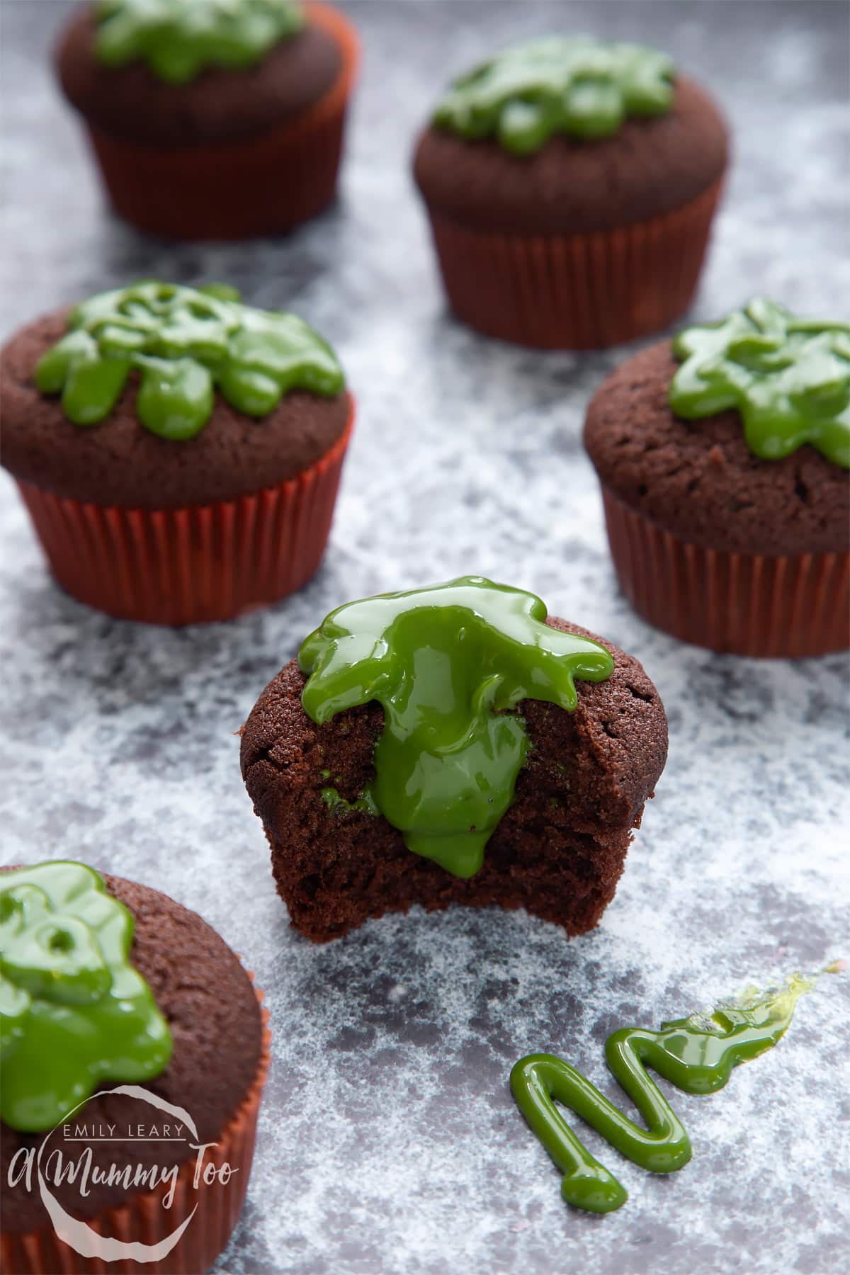 Slime cupcakes on a black backdrop. The cakes have a chocolate sponge topped with dyed-green caramel. The cupcake in the foreground has been bitten into, revealing the caramel core.