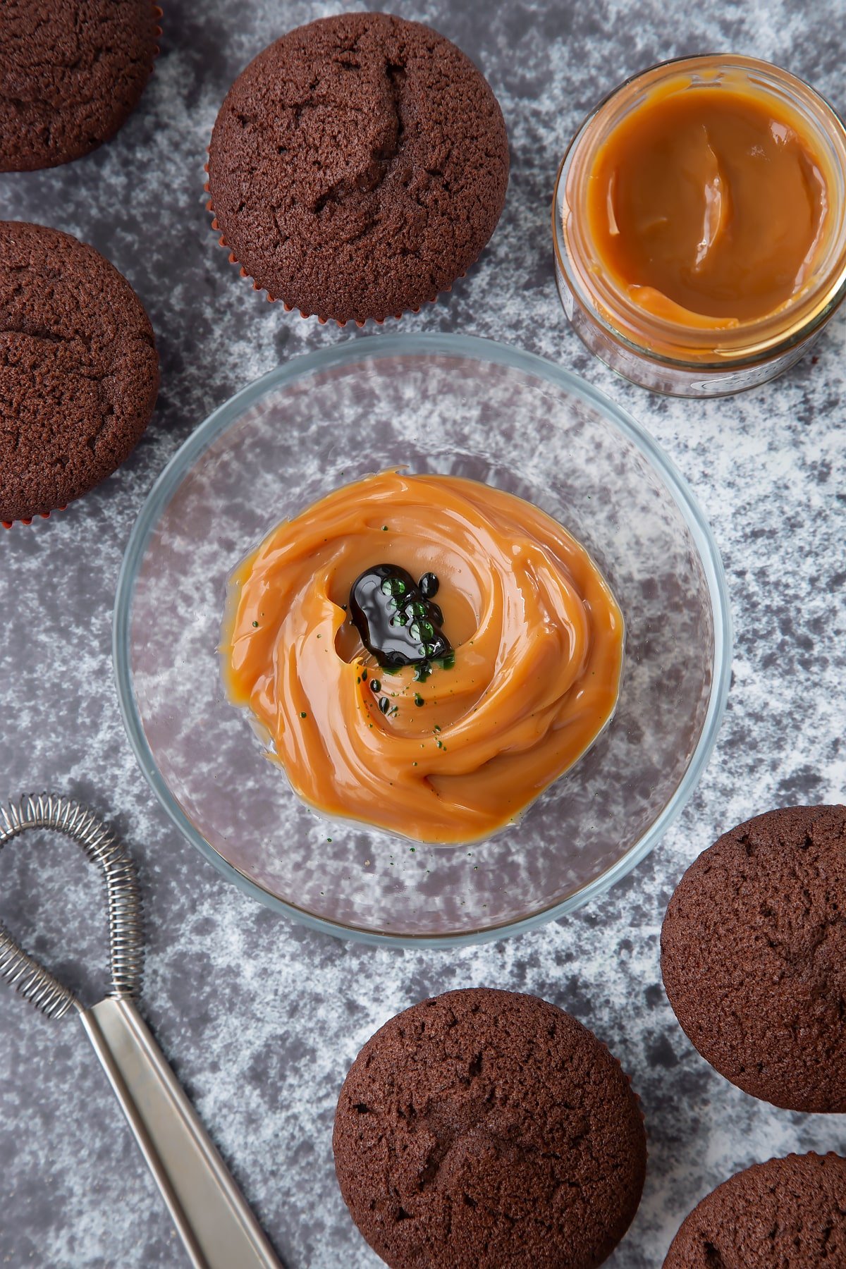 A small glass bowl of caramel topped with green food colouring. Chocolate cupcakes surround the bowl.