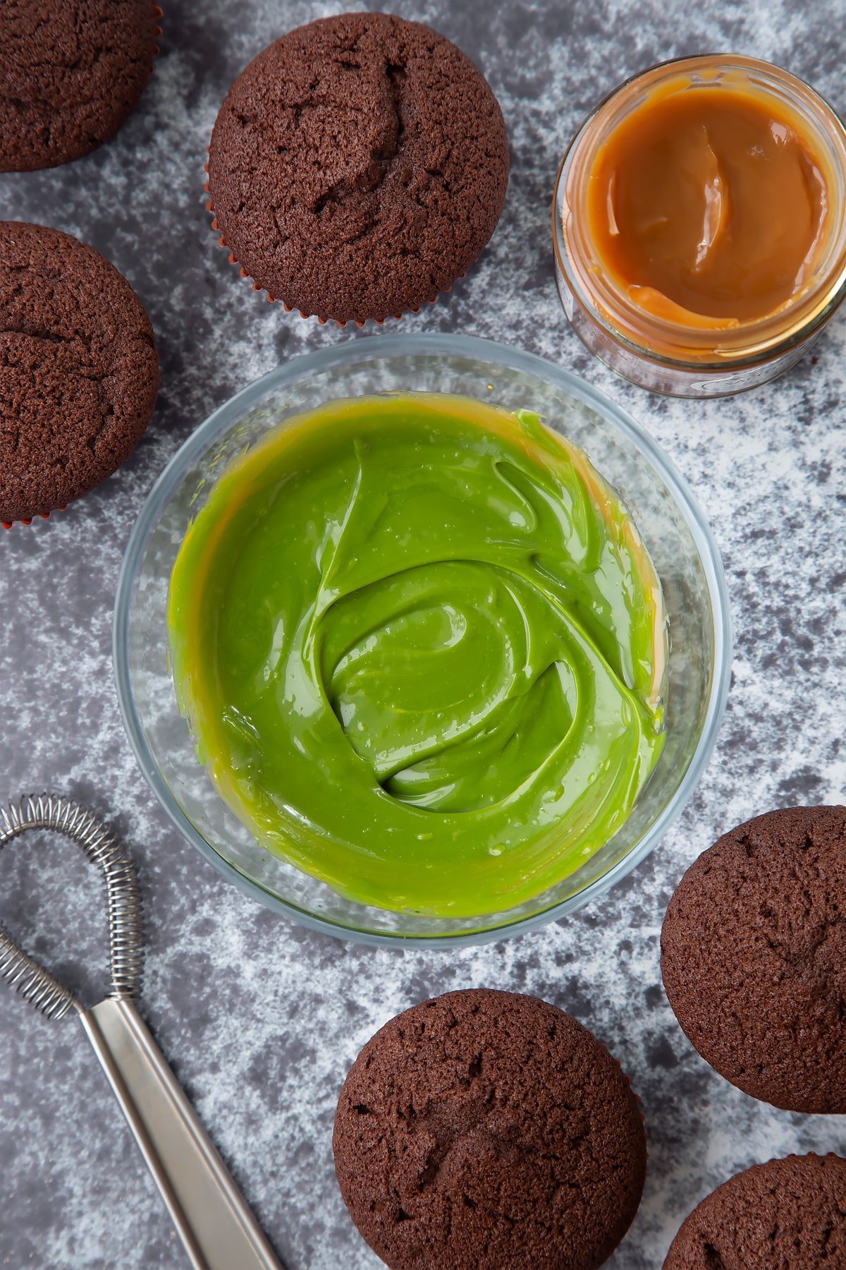 A small glass bowl of  dyed-green caramel. Chocolate cupcakes surround the bowl.