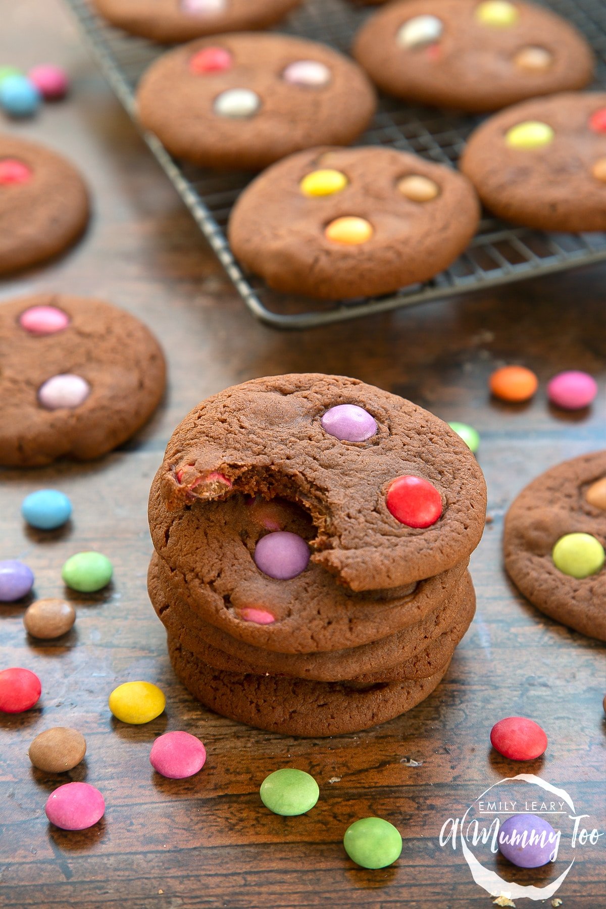 Smartie cookies stacked up on a dark wooden surface. The top cookie has been bitten.