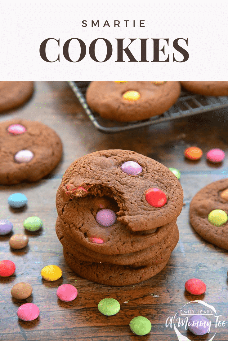 Smartie cookies stacked up on a dark wooden surface. The top cookie has been bitten. Caption reads: Smartie cookies