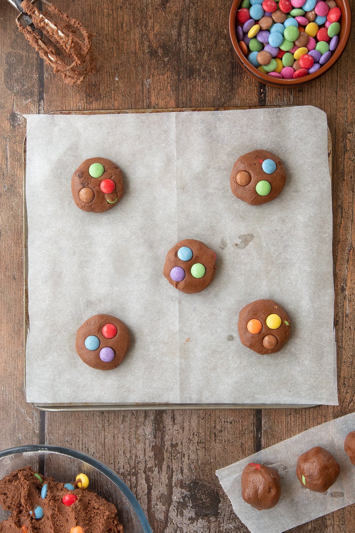 Smartie cookies on a baking tray lined with baking paper, ready to bake. Ingredients to make the Smartie cookie recipe surround the tray. 