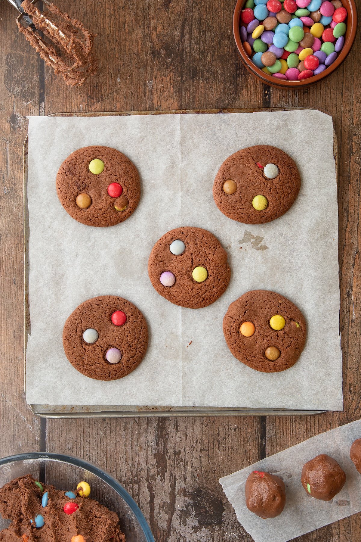 Freshly baked Smartie cookies on a baking tray lined with baking paper. Ingredients to make the Smartie cookie recipe surround the tray. 