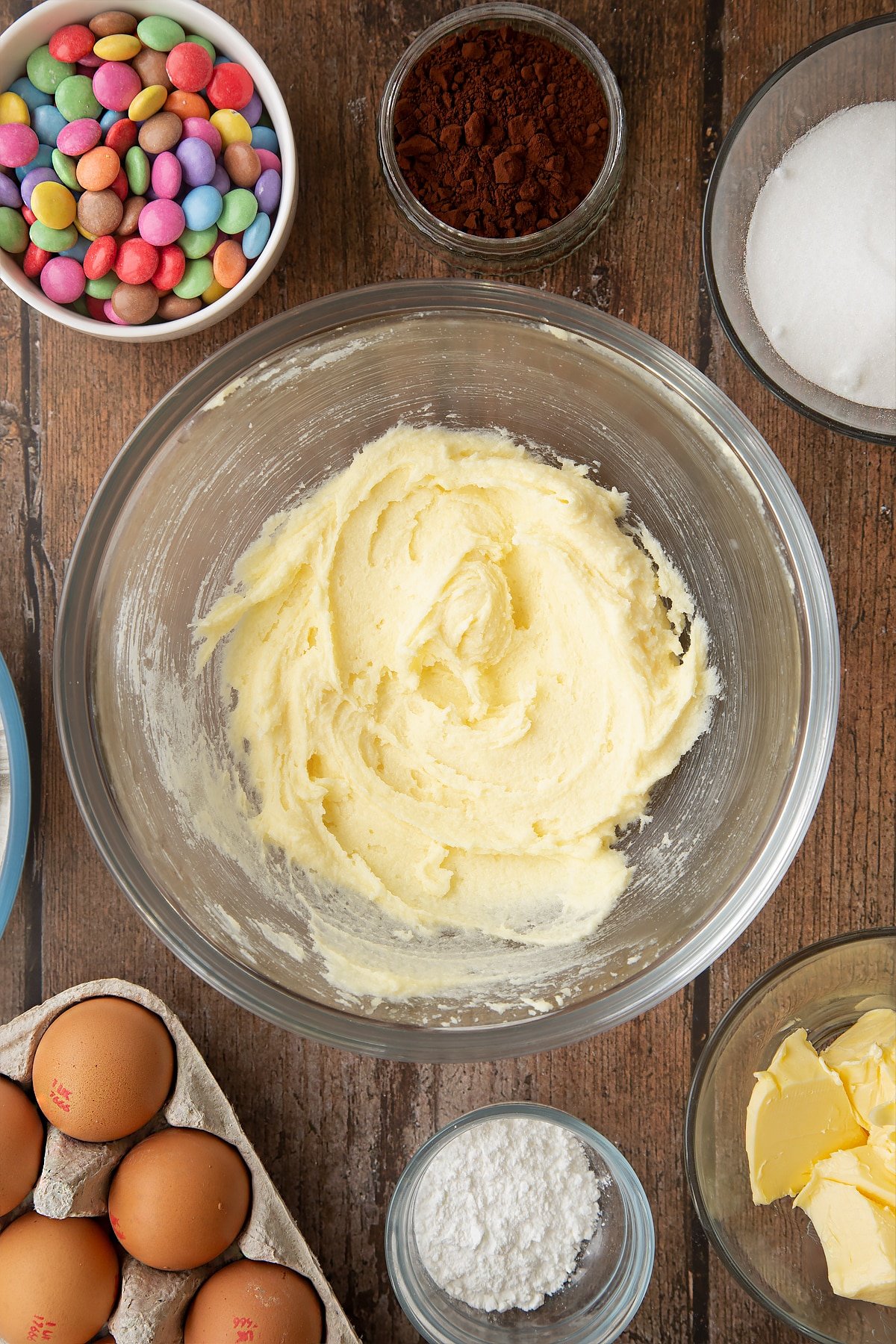 Beaten sugar and butter in a glass mixing bowl. Ingredients to make the Smartie cookie recipe surround the bowl. 