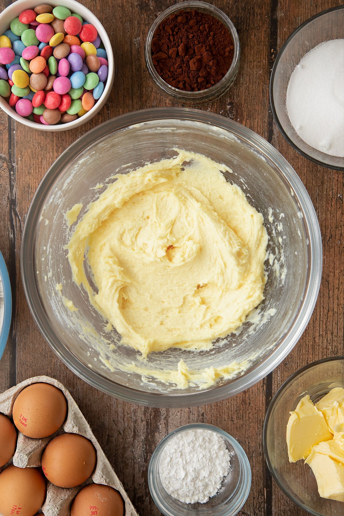 Beaten sugar, eggs and butter in a glass mixing bowl. Ingredients to make the Smartie cookie recipe surround the bowl. 