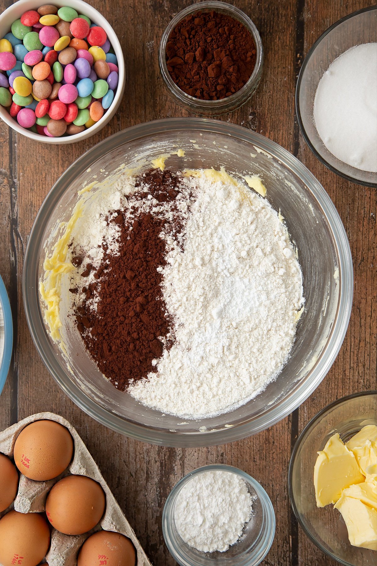 Beaten sugar, eggs and butter, topped with flour, cocoa and baking powder in a glass mixing bowl. Ingredients to make the Smartie cookie recipe surround the bowl. 