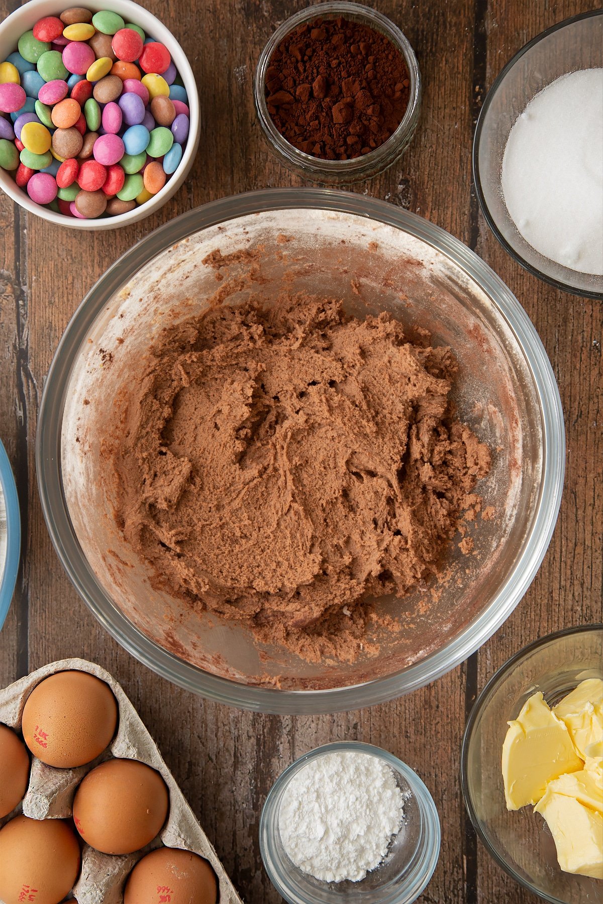 Chocolate cookie dough in a glass mixing bowl. Ingredients to make the Smartie cookie recipe surround the bowl. 