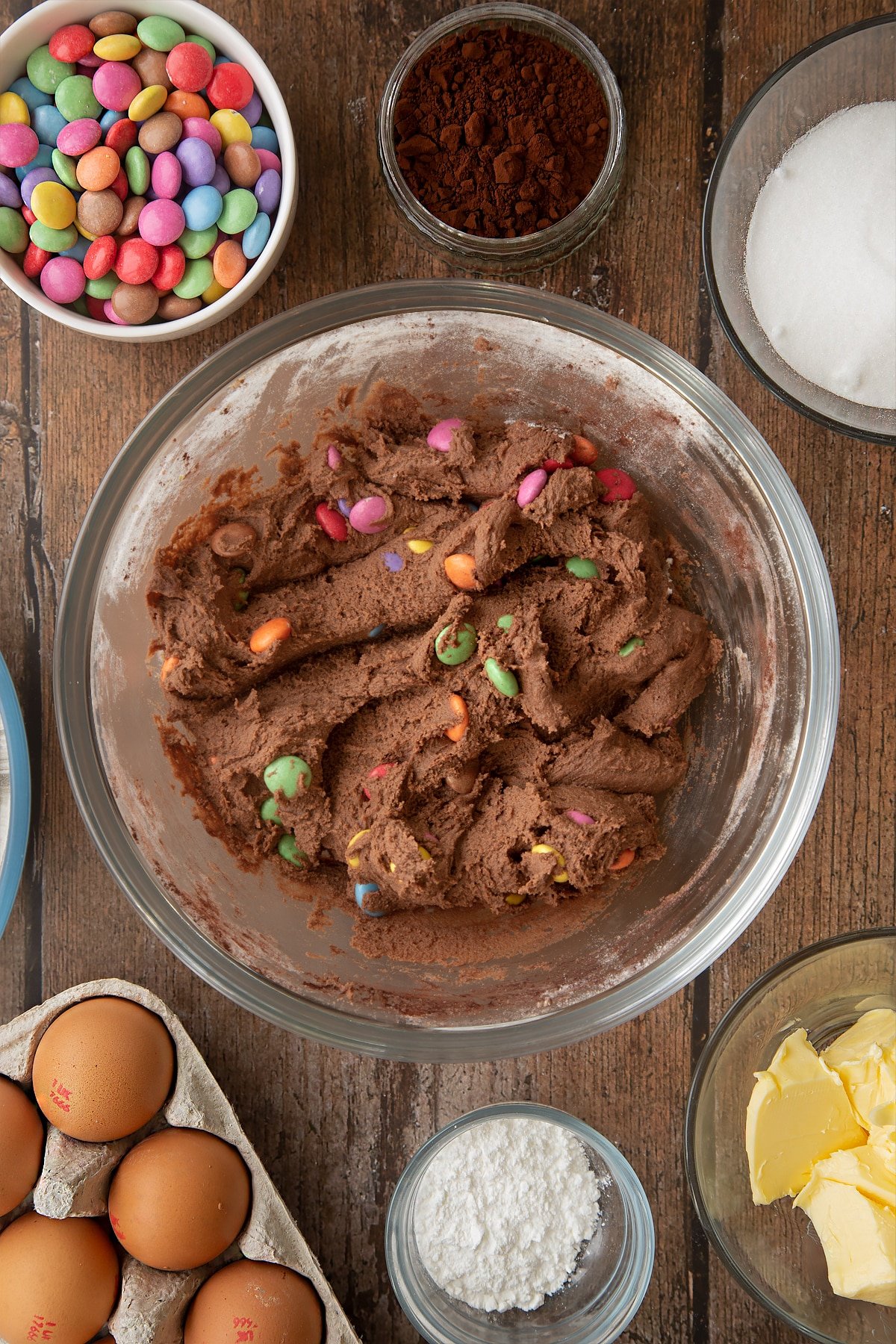 Smartie cookie dough in a glass mixing bowl. Ingredients to make the Smartie cookie recipe surround the bowl. 