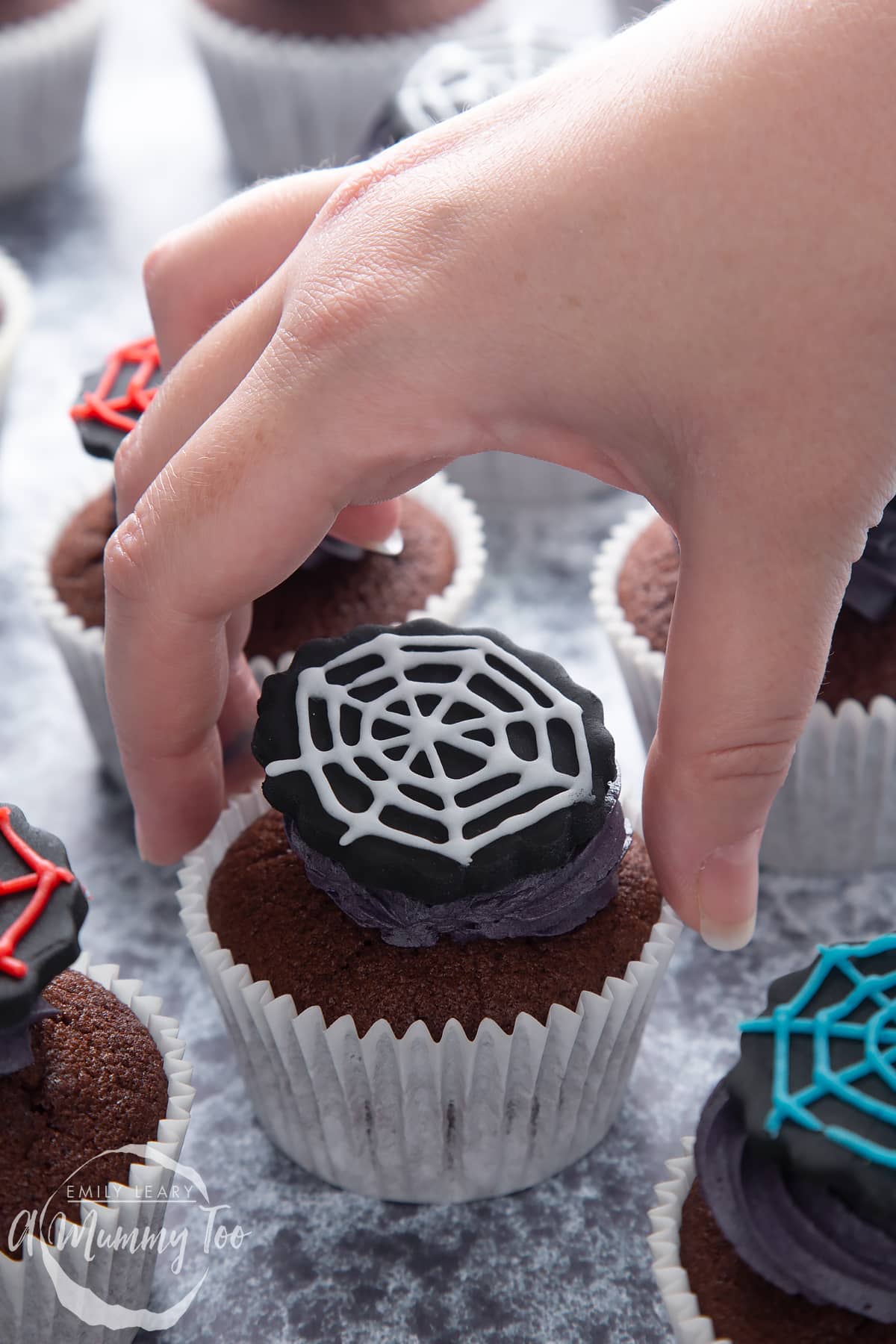 Spider web cupcakes arranged on a black surface. The chocolate sponge cupcakes are topped with purple vanilla buttercream and discs of black sugar paste decorated with spider web icing. A hand reaches to take one.