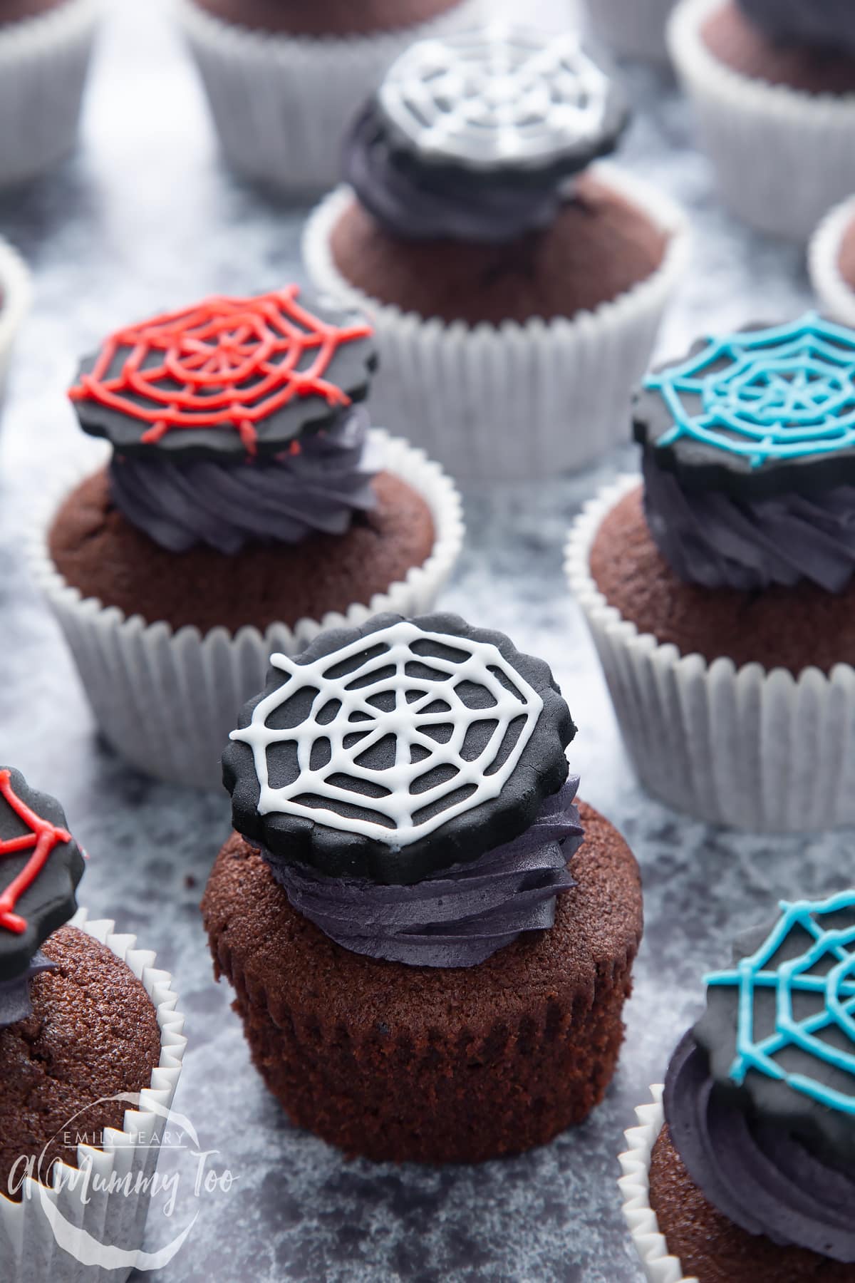 Spider web cupcakes arranged on a black surface. The chocolate sponge cupcakes are topped with purple vanilla buttercream and discs of black sugar paste decorated with spider web icing.