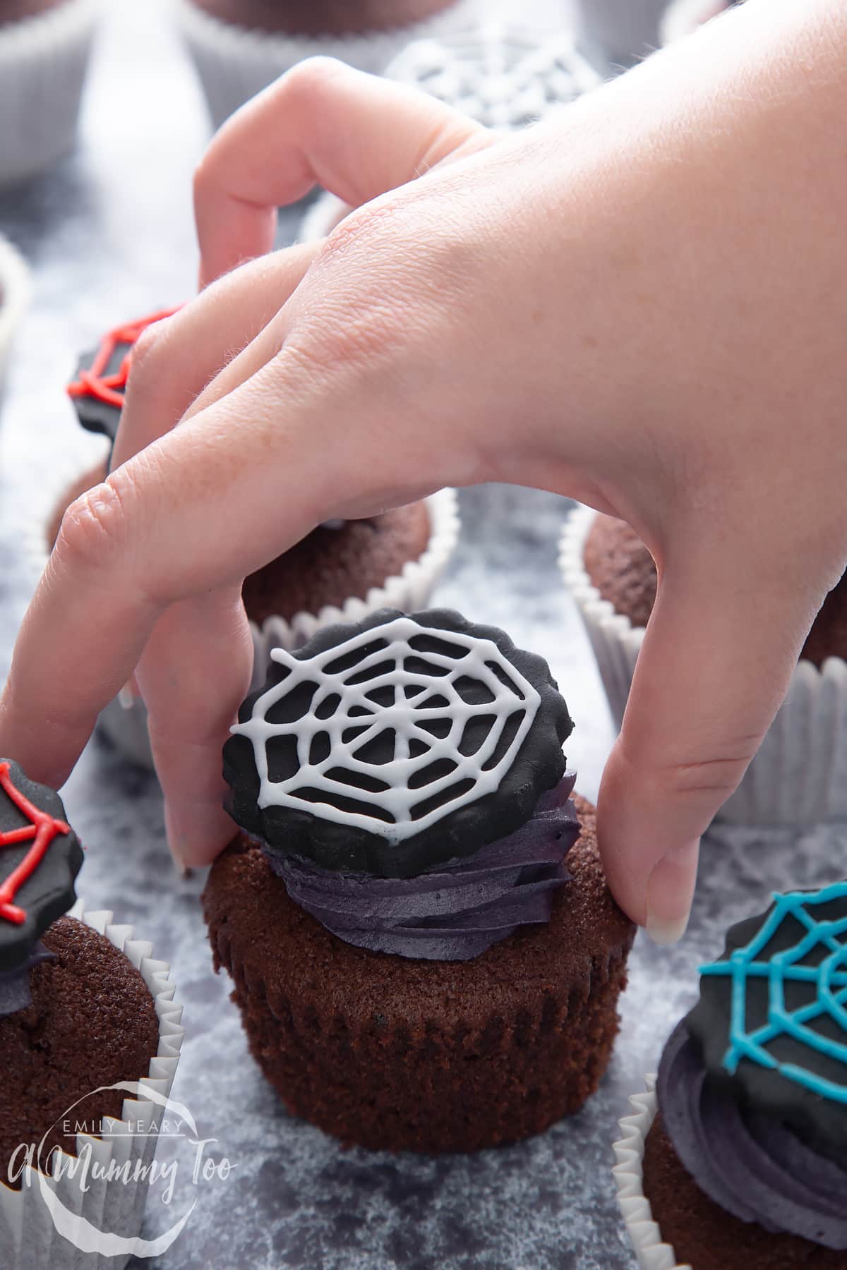 Spider web cupcakes arranged on a black surface. The chocolate sponge cupcakes are topped with purple vanilla buttercream and discs of black sugar paste decorated with spider web icing. A hand reaches to take an unwrapped one.