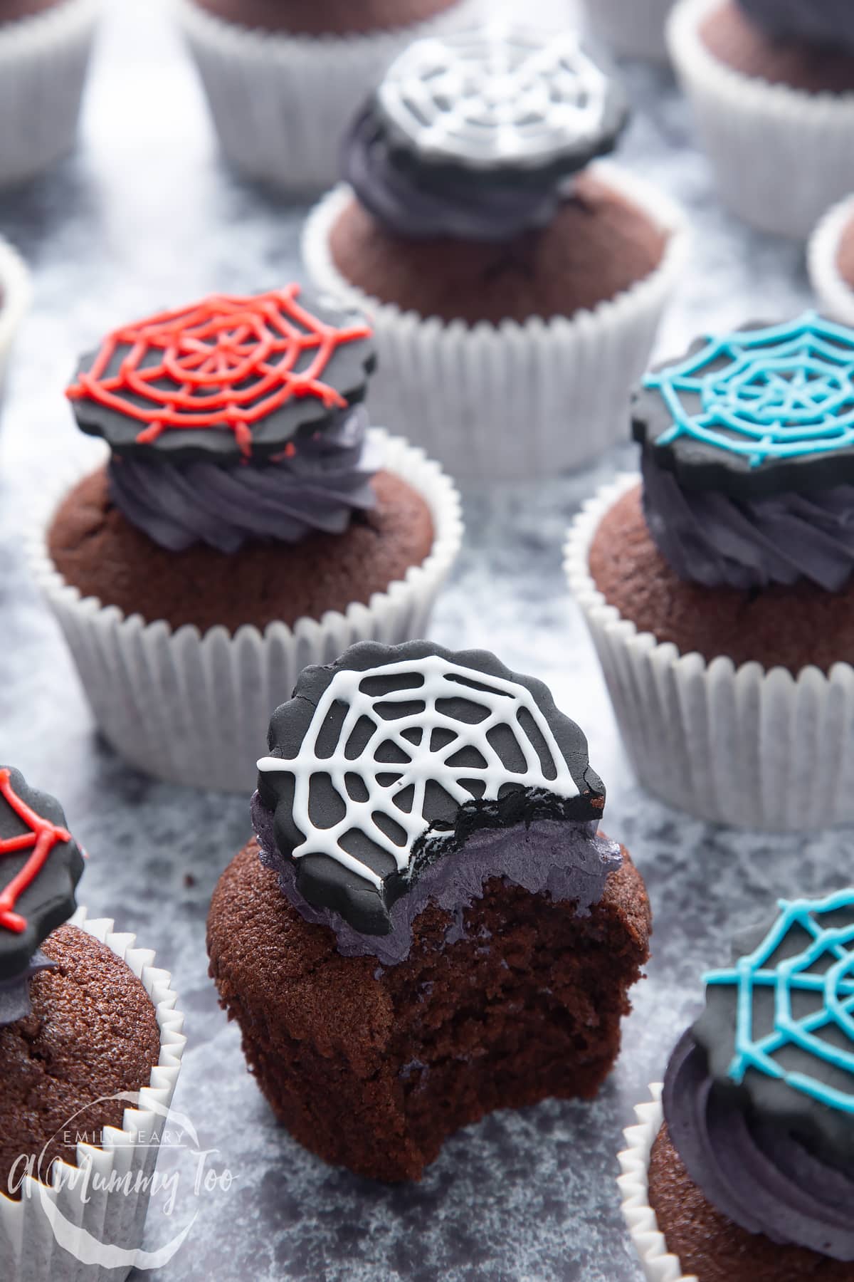 Spider web cupcakes arranged on a black surface. The chocolate sponge cupcakes are topped with purple vanilla buttercream and discs of black sugar paste decorated with spider web icing. One cupcake has been bitten.