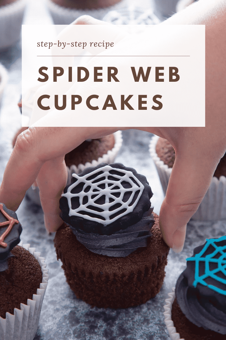 Spider web cupcakes arranged on a black surface. The chocolate sponge cupcakes are topped with purple vanilla buttercream and discs of black sugar paste decorated with spider web icing. A hand reaches for one. Caption reads: Step-by-step recipe spider web cupcakes