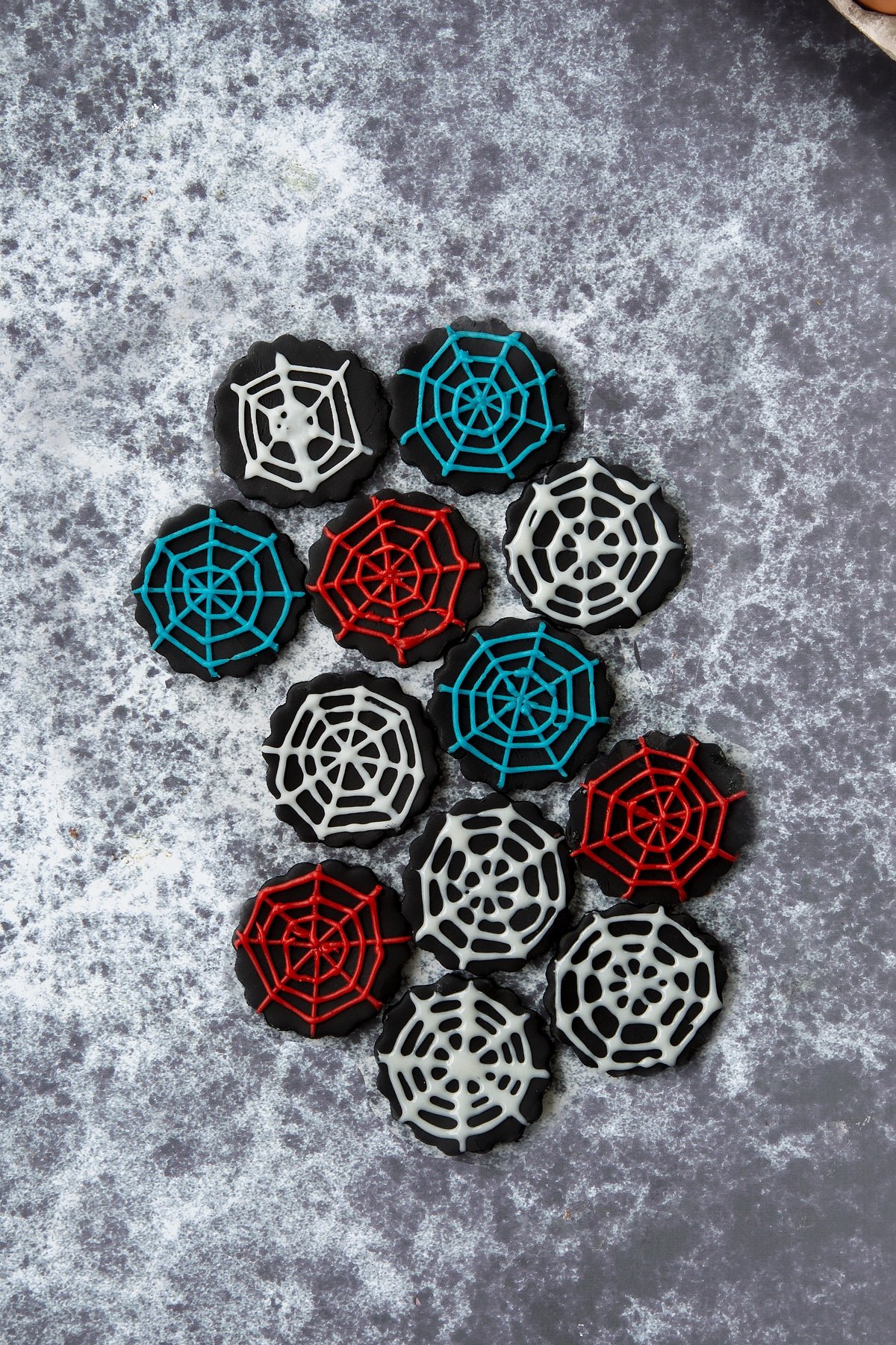 Discs of black sugar paste on a dark background. The discs have been decorated with spider webs using red, blue and white icing pens.