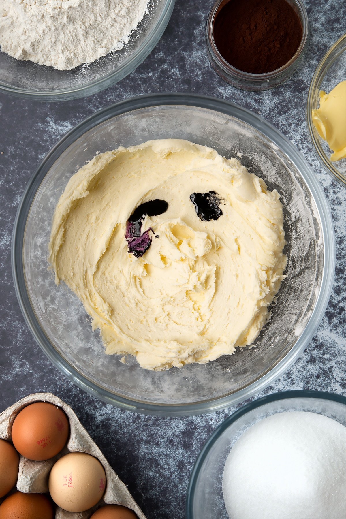 Icing sugar, vanilla and butter whisked together in a large mixing bowl. Purple and black food colouring has been added. Ingredients to make spider web cupcakes surround the bowl.