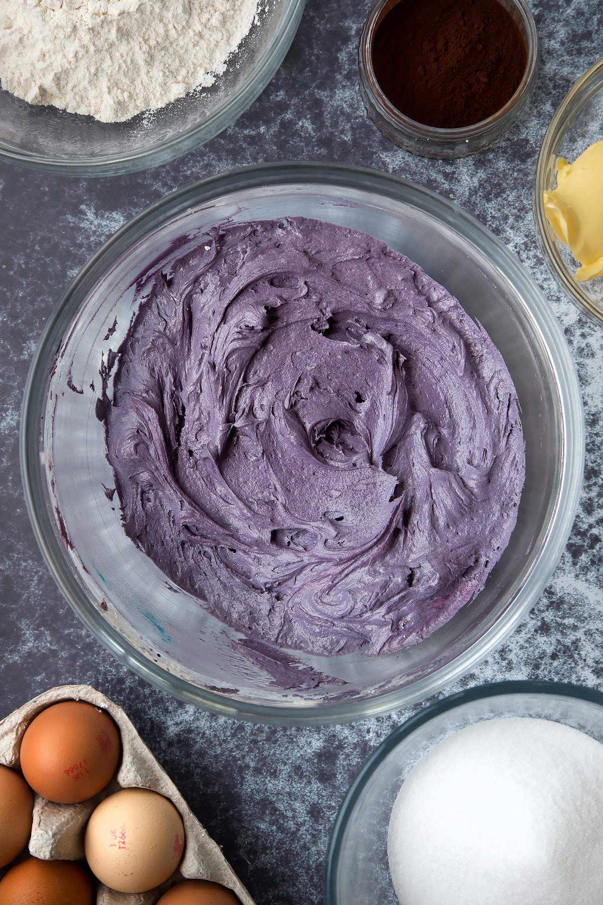 Purple buttercream in a large mixing bowl. Ingredients to make spider web cupcakes surround the bowl.