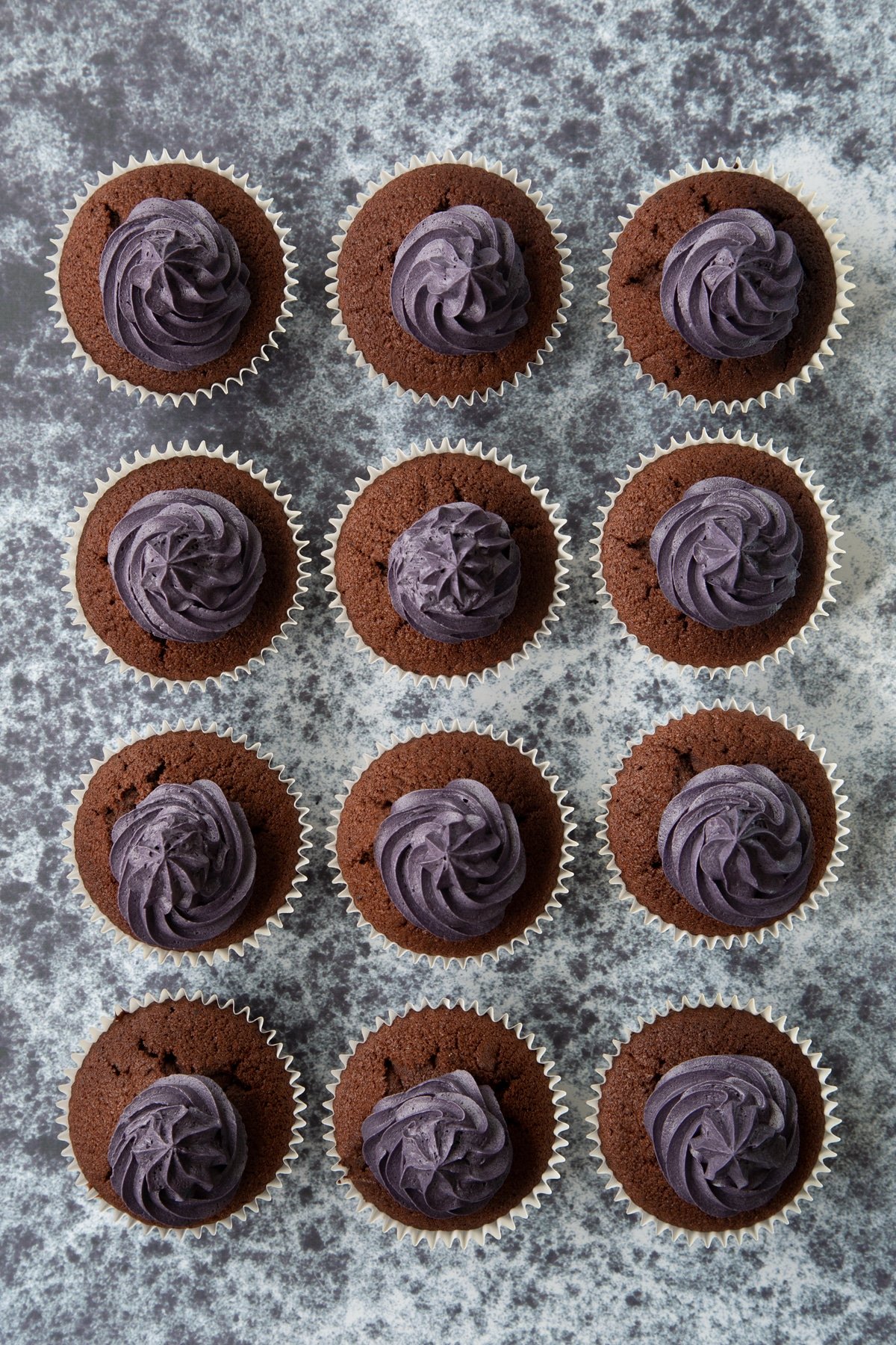 12 chocolate cupcakes with purple frosting on a dark background, shown from above.