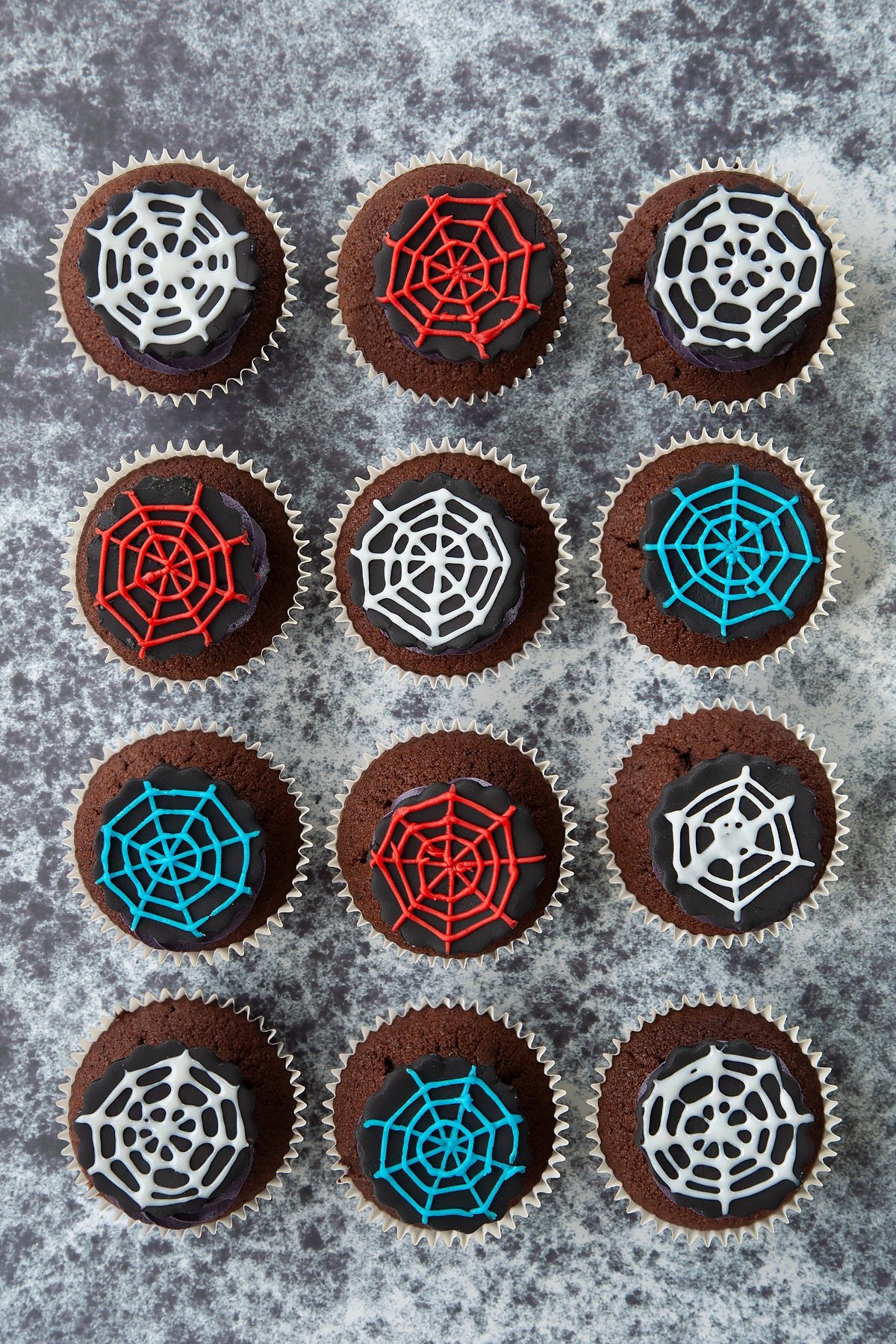 Spider web cupcakes arranged on a black surface, shown from above. The chocolate sponge cupcakes are topped with purple vanilla buttercream and discs of black sugar paste decorated with spider web icing.