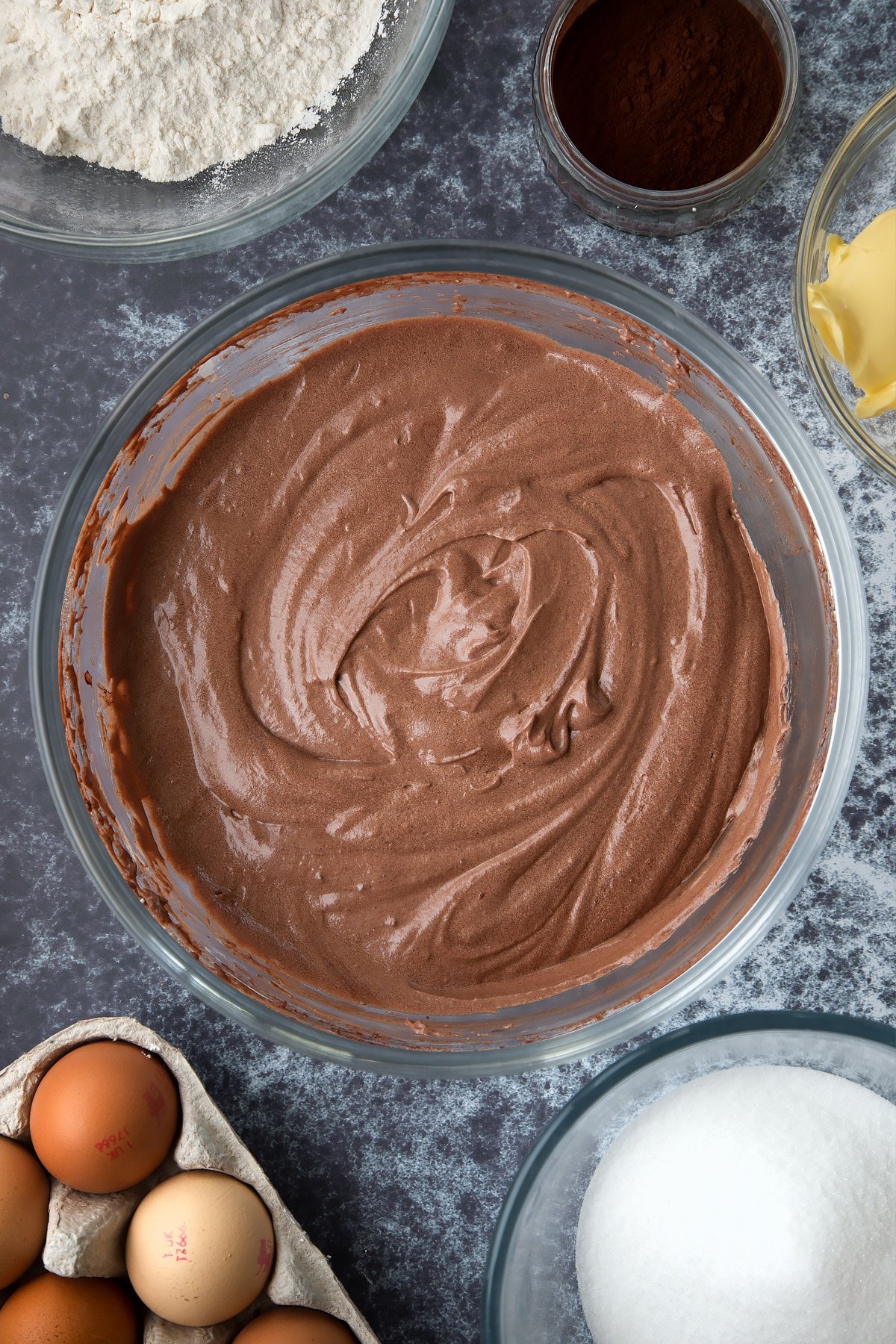 Chocolate cupcake batter in a large mixing bowl. Ingredients to make spider web cupcakes surround the bowl.