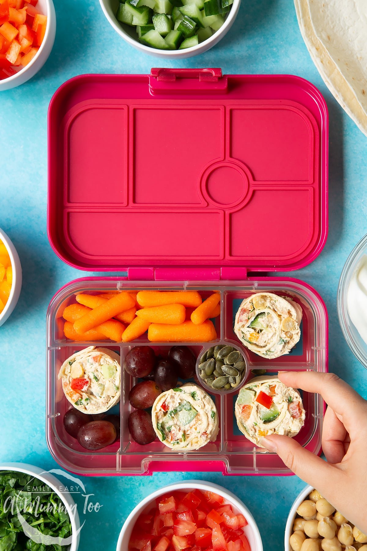 Tuna pinwheels in a lunchbox with fresh fruit and veg. A hand reaches to take a pinwheel.