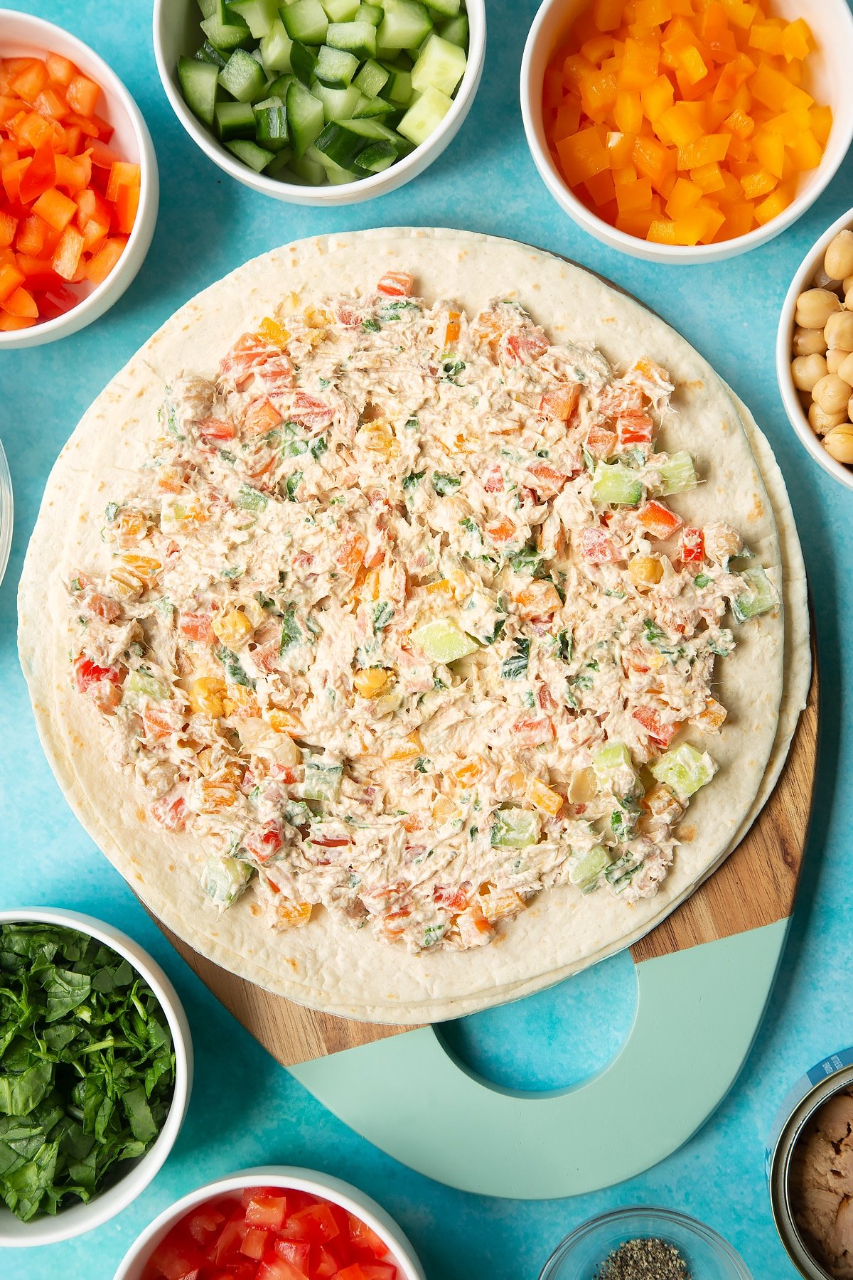 Flour tortillas spread with a mix of cream cheese, tuna and veg. Ingredients to make tuna pinwheels surround the board.