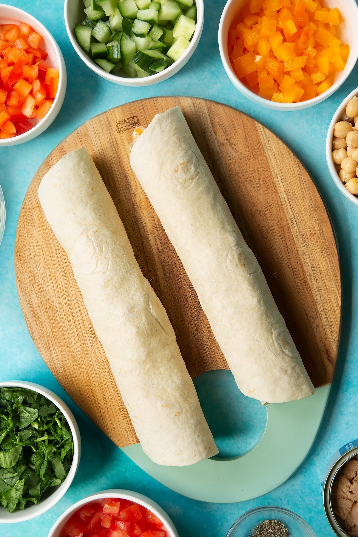 Two rolled up flour tortillas on a wooden board. Ingredients to make tuna pinwheels surround the board.