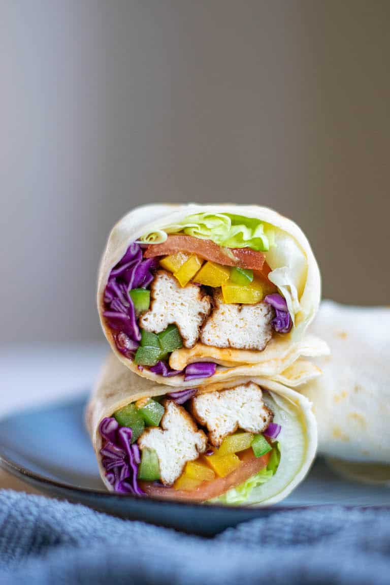 Tofu wrap with red cabage, lettuace and tomatoes sits on a black plate. The wrap is cut open so you can see the contents inside which are facing the camera.