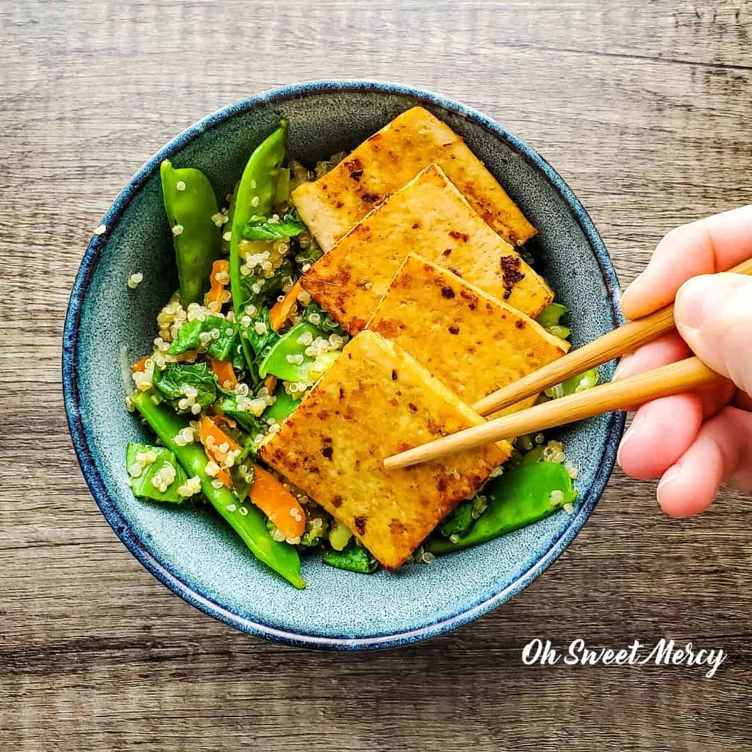 Slices of tofu in a decorative bowl with garlic, ginger, slices of lime, quinoa and green beans. A hand reaches in with a pair of chopsticks to pick a slice of the tofu up.