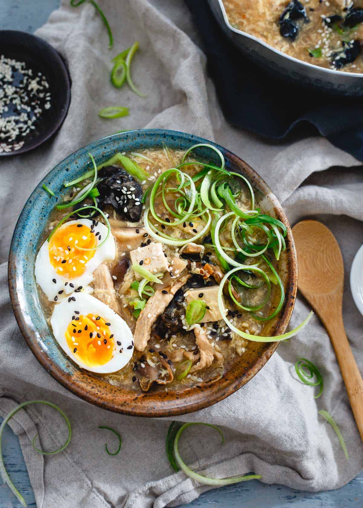 Bowl of hot and sour egg drop soup made from tofu on top of a table cloth with a bowl of sesame seeds at the side. 